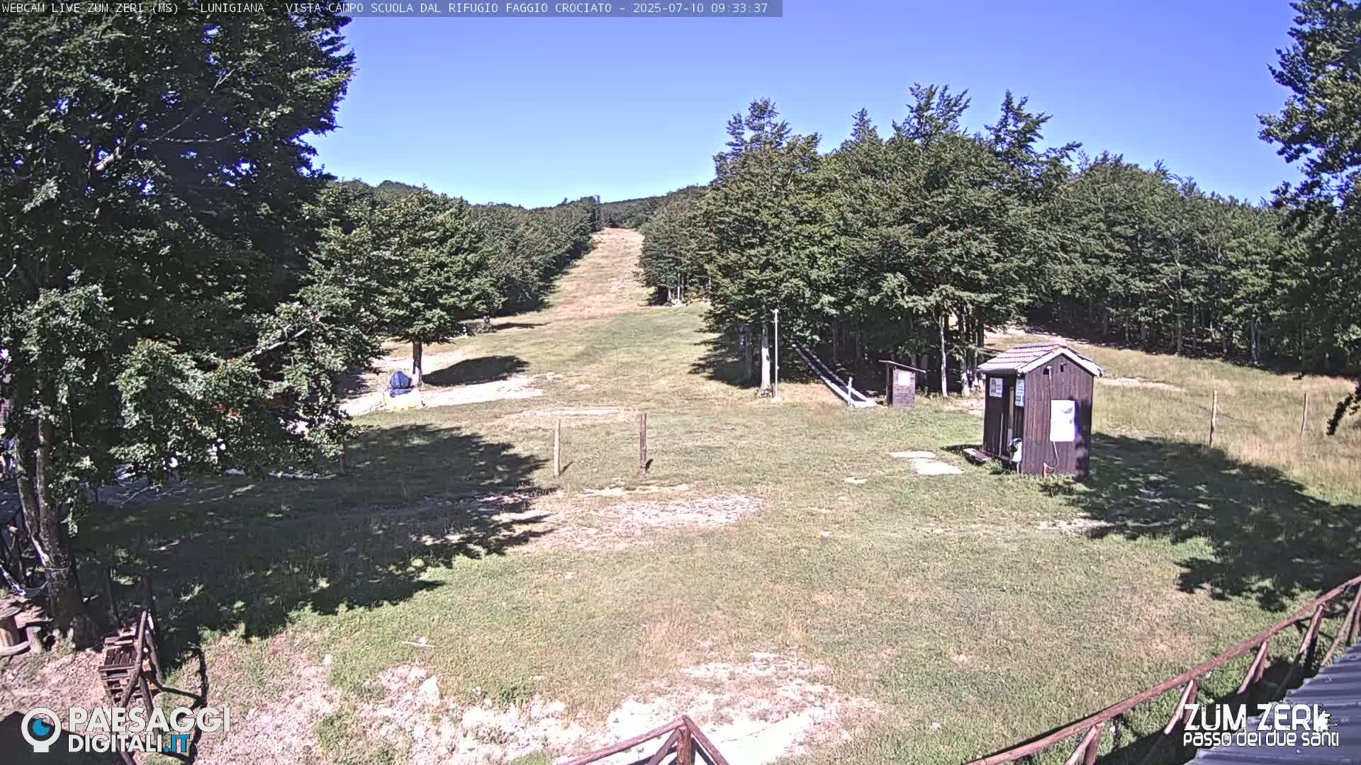 A grassy clearing with a small shed and a ski slope is visible under a clear blue sky, surrounded by lush green trees.