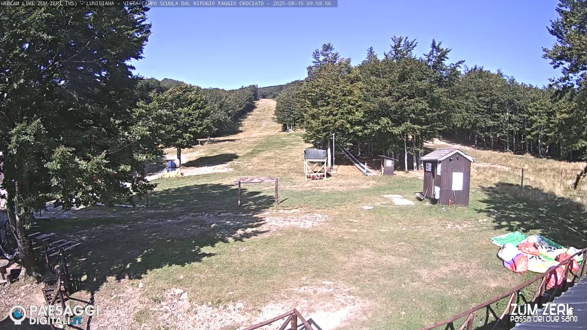 A grassy clearing with a small building and a ski slope is visible under a sunny sky, surrounded by trees.