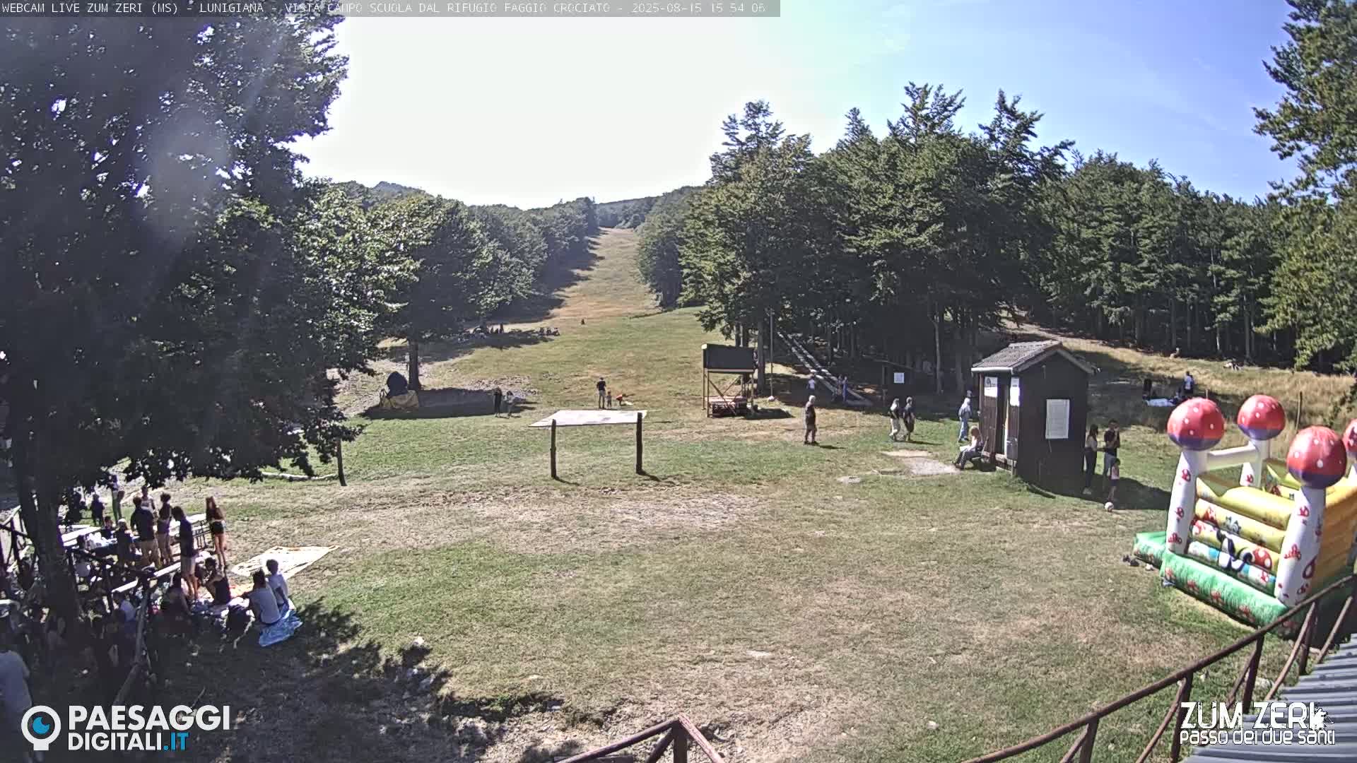 A grassy clearing with a small building and a ski slope is visible under a sunny sky, surrounded by trees.