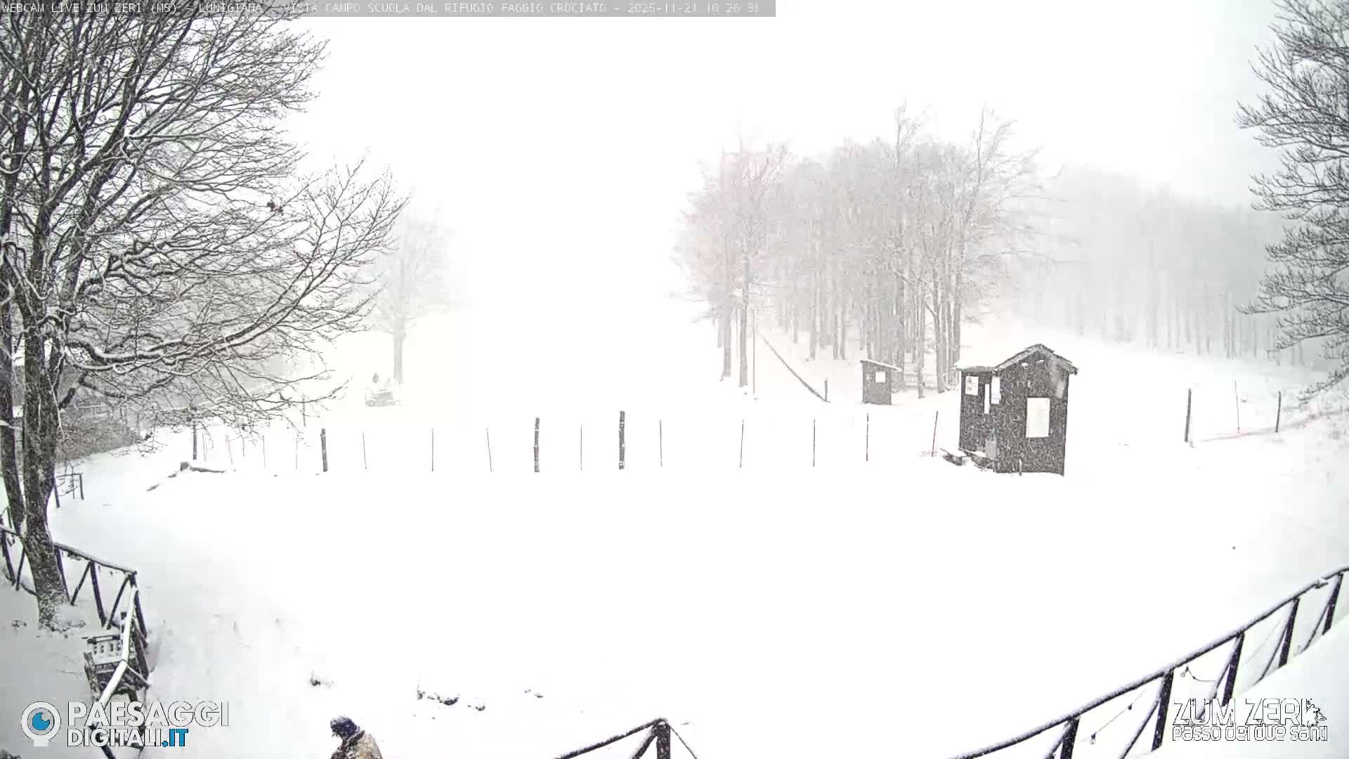 A vast, snow-covered winter landscape features bare trees with snow on their branches, two small dark huts, and a fence line, all obscured by heavy snowfall and low visibility.