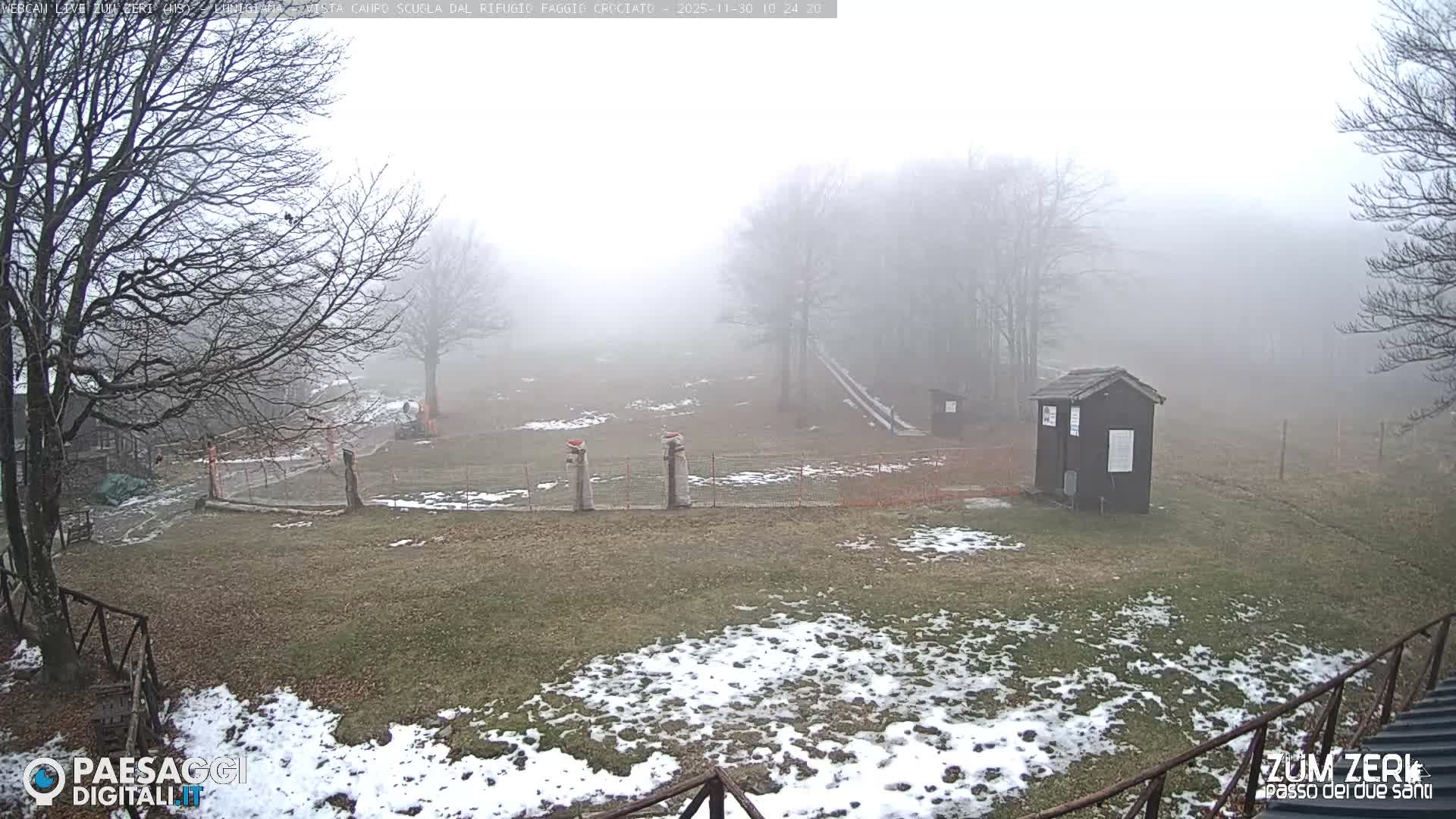 A very foggy outdoor scene displays a winter landscape with bare trees, patches of snow on sparse grassy ground, and a small dark wooden hut in the foreground.