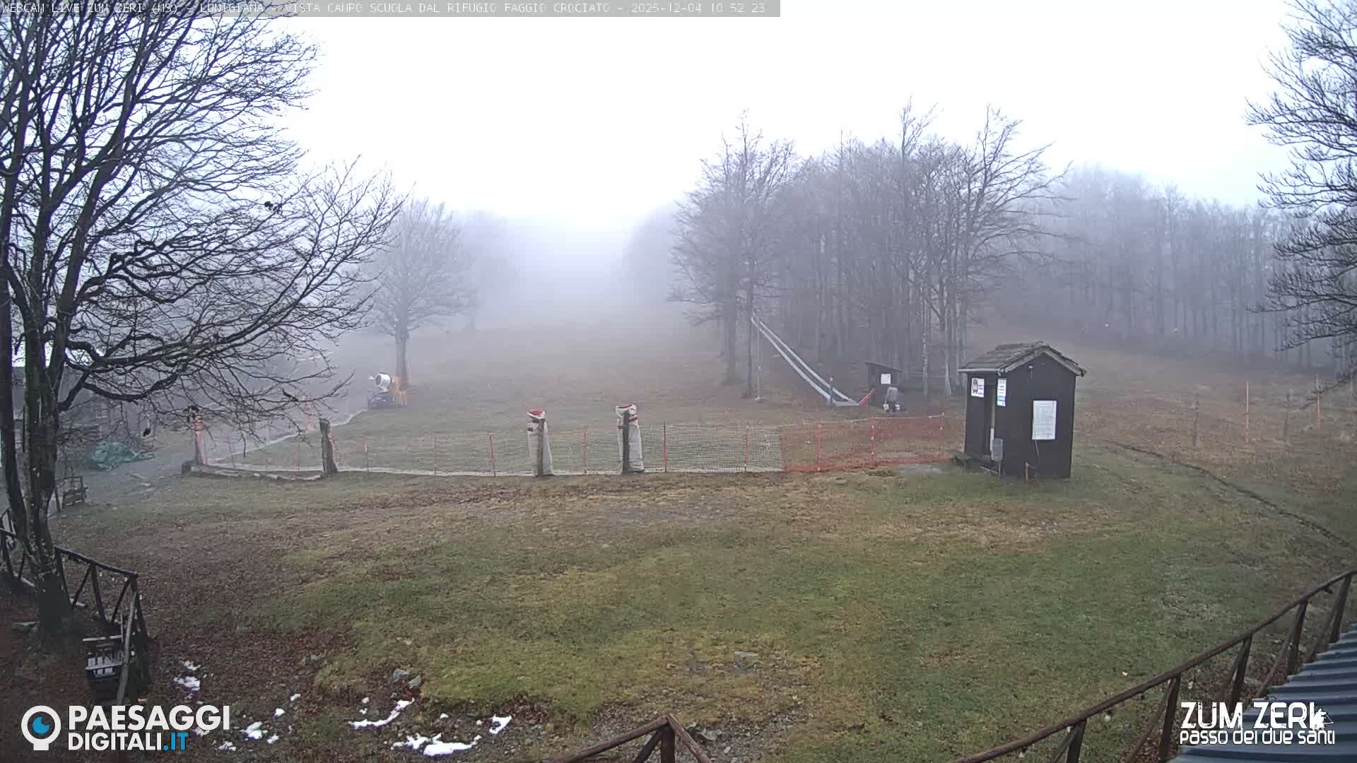 A misty winter landscape features bare trees, a small wooden hut, and ski infrastructure on brown and green ground with patches of snow, all shrouded in dense fog.