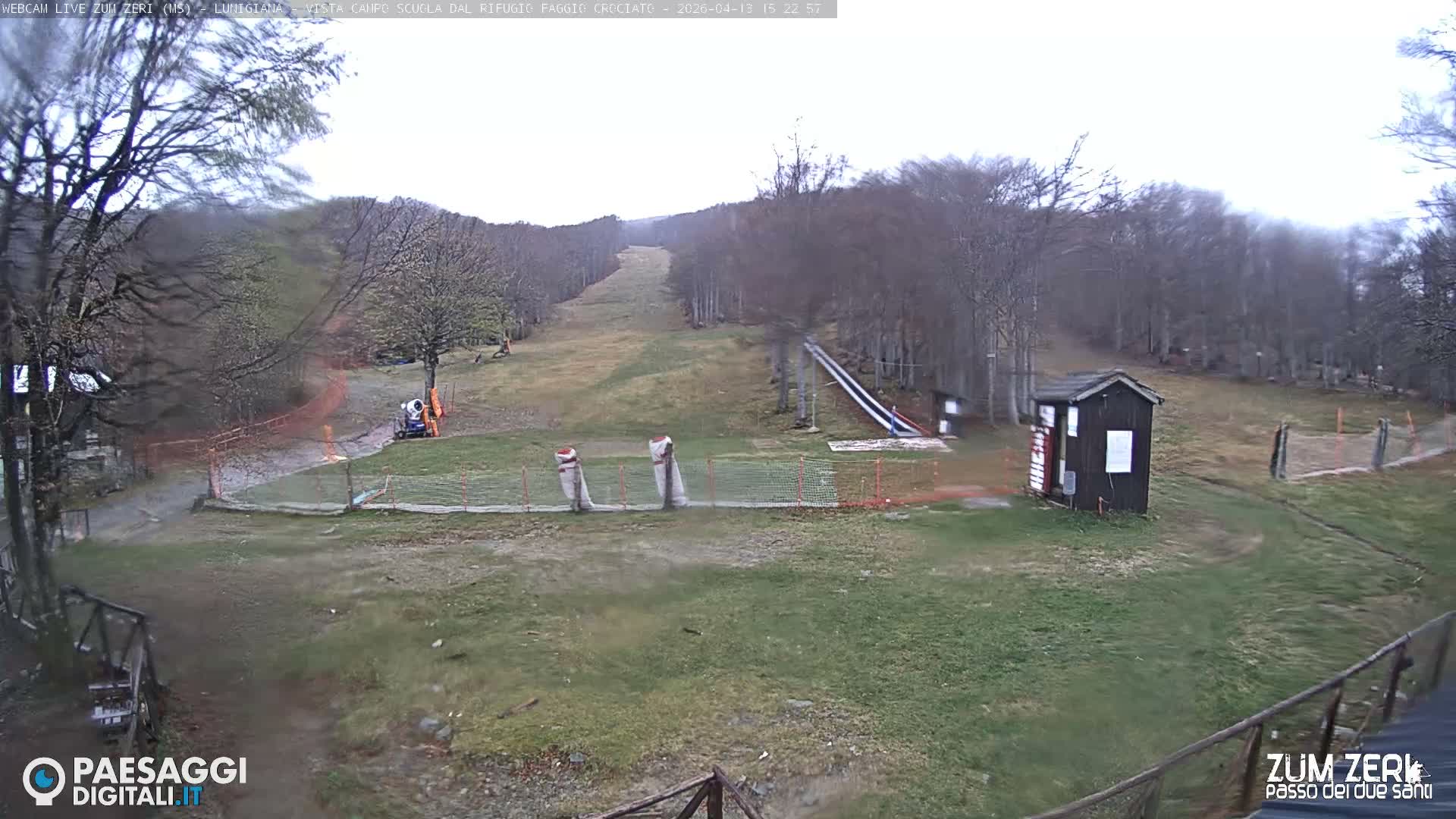 A misty winter landscape features bare trees, a small wooden hut, and ski infrastructure on brown and green ground with patches of snow, all shrouded in dense fog.