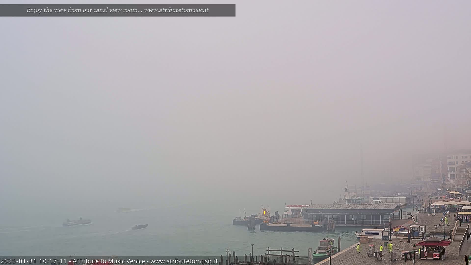 A foggy view of a canal in Venice shows several boats and a dock with people near buildings in the distance.