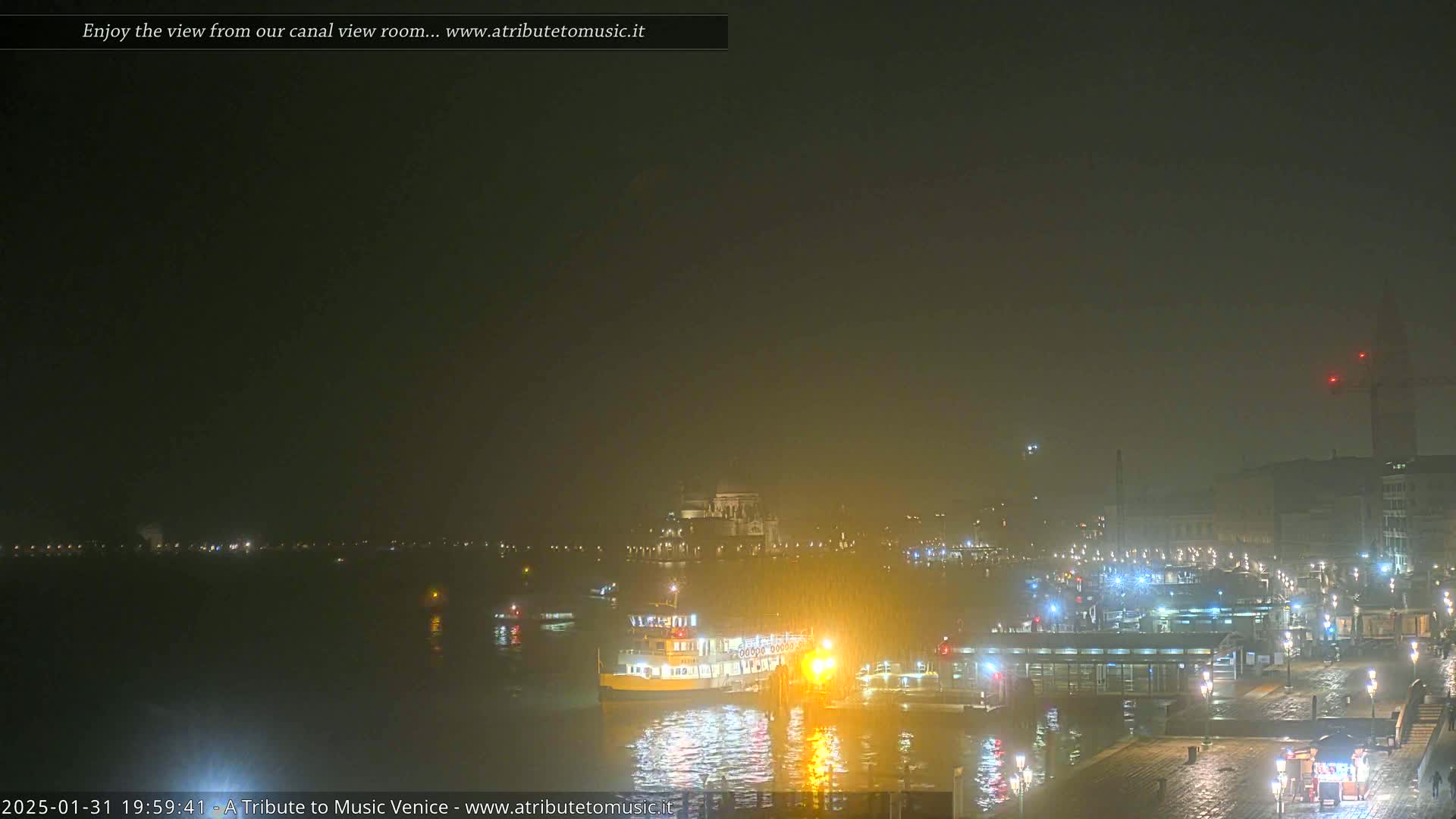 A nighttime view of a waterway with a boat docked near a city under hazy, possibly foggy conditions.