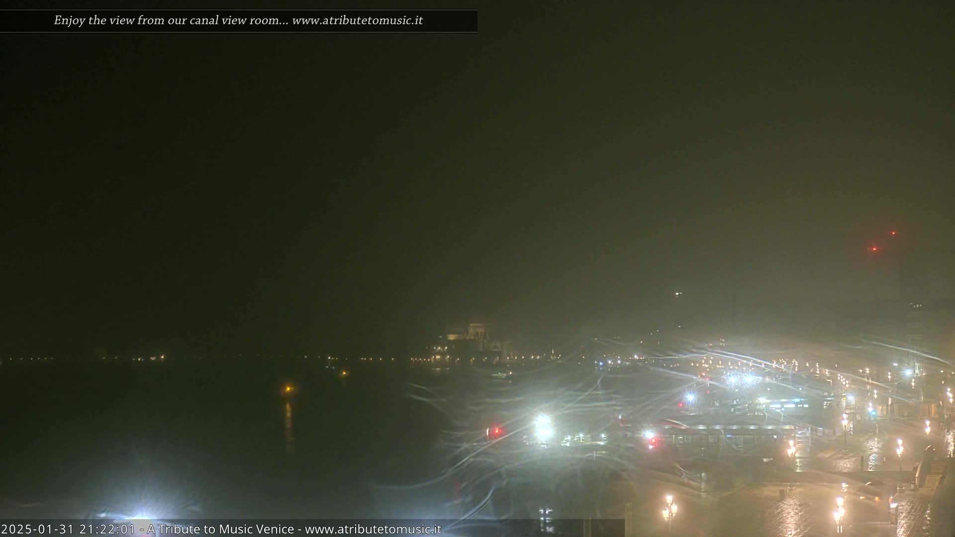 A nighttime view of a canal and city waterfront in snowy or windy conditions, showing blurry lights and buildings across the water.