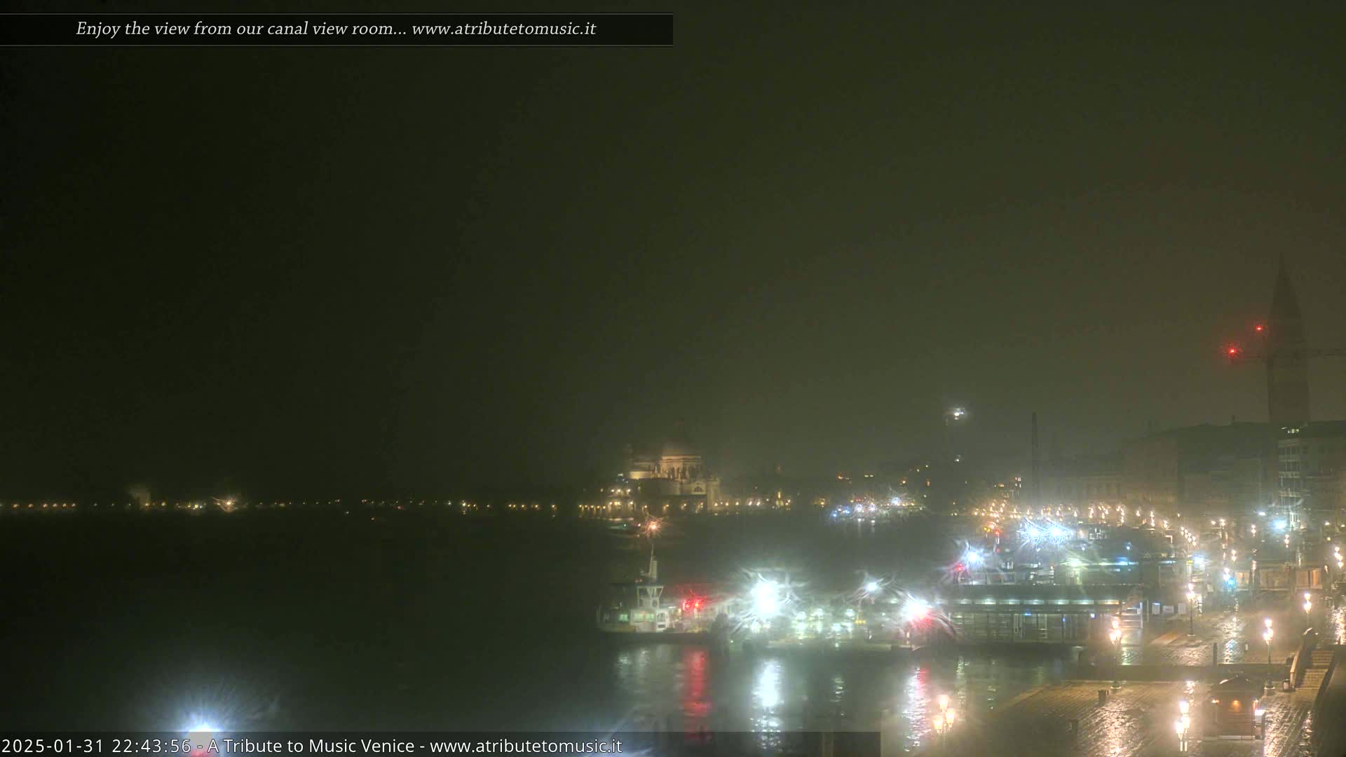 A nighttime view of a canal, showing dimly lit buildings, boats, and a large building with a dome in the distance, under hazy or foggy conditions.