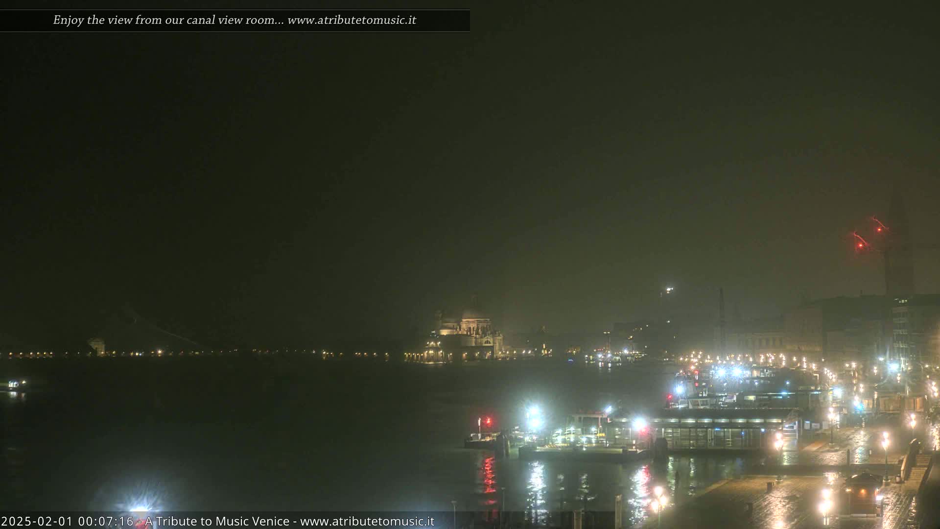 A nighttime view of a canal, with a large building faintly visible in the distance under hazy conditions, shows several boats docked at a waterfront area.
