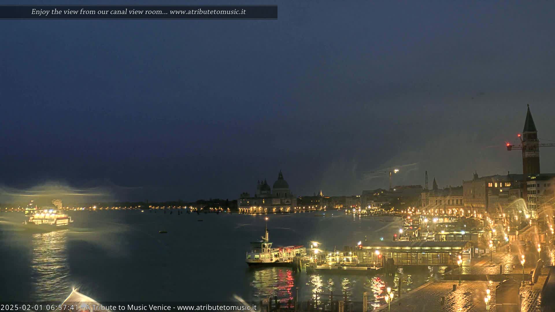 A nighttime view of a canal in Venice, Italy, shows illuminated buildings, boats, and a water taxi, with a slightly misty or rainy atmosphere.