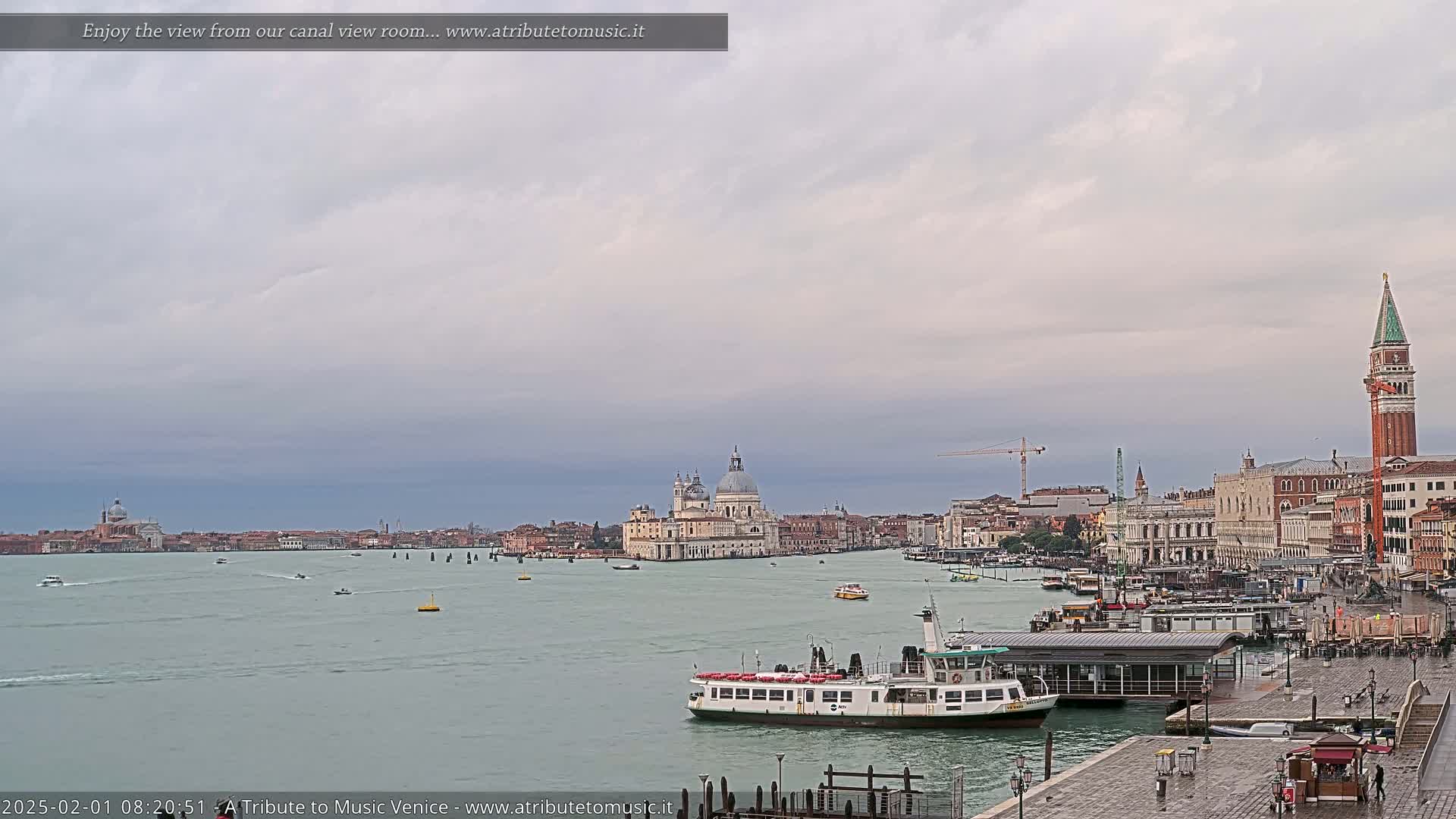 A wide shot of a Venetian canal on an overcast day shows a ferry boat near a dock, with the Basilica di Santa Maria della Salute and St. Mark's Campanile in the distance.