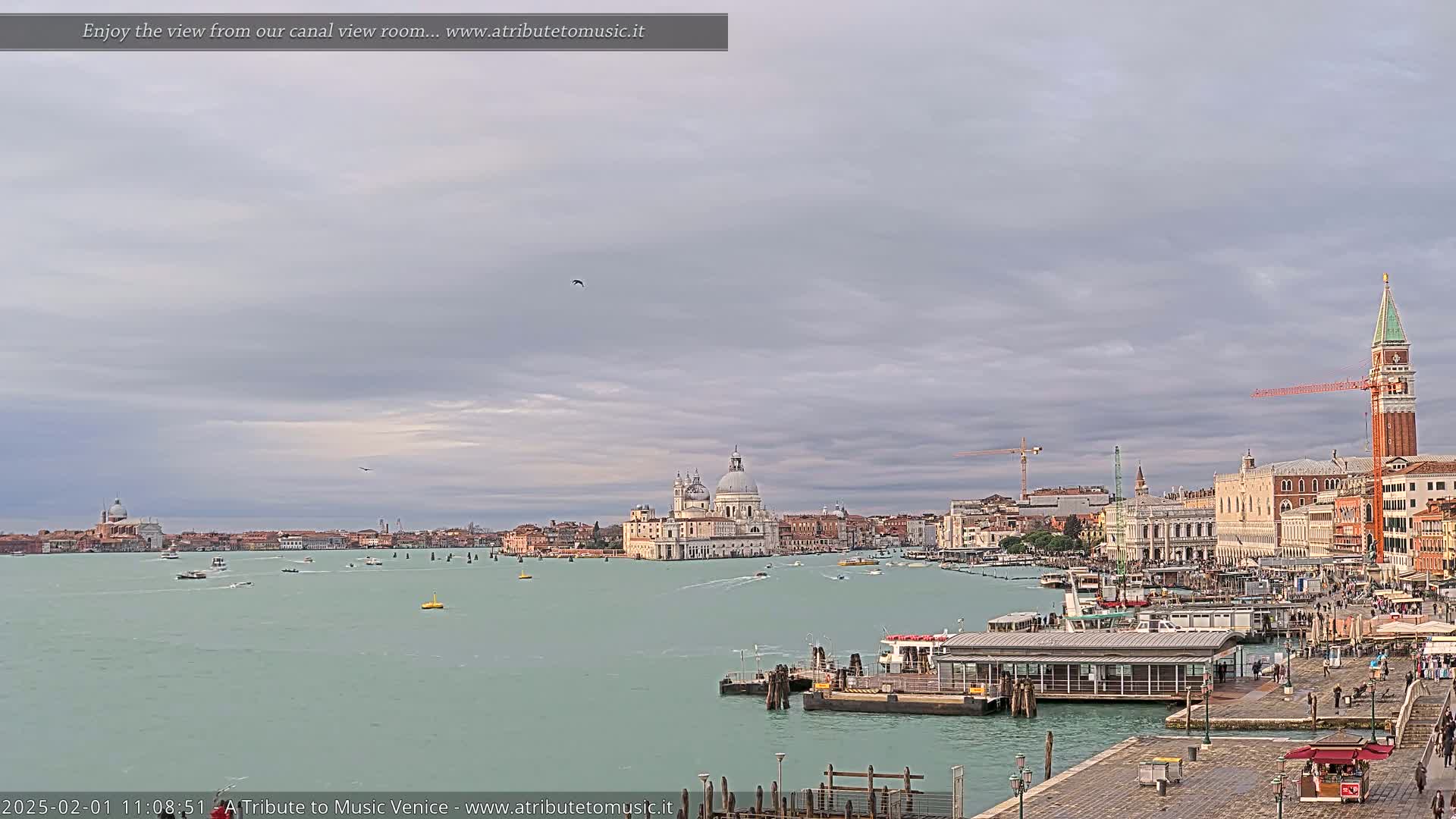 A wide view of a Venetian canal on an overcast day shows boats on the water, buildings along the shore, and a large church in the distance.