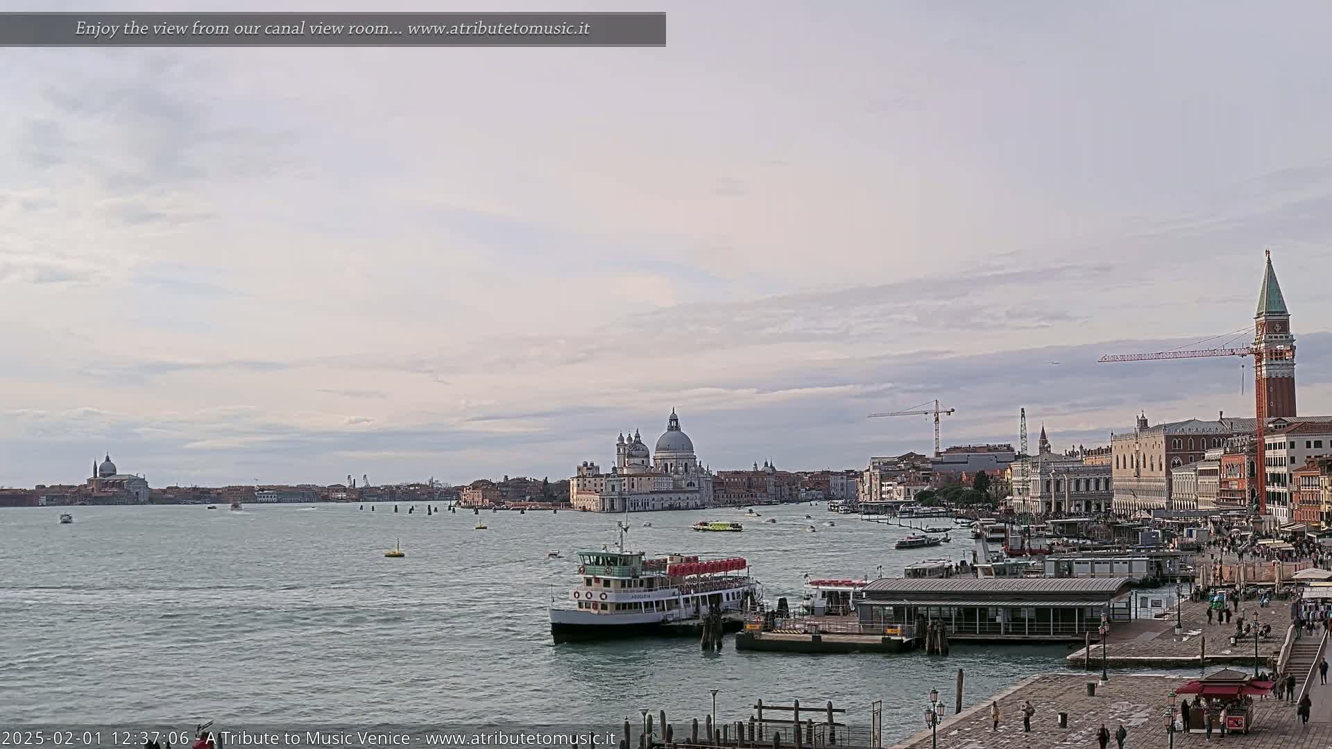 A wide, calm canal scene in Venice shows several boats, a large building with a dome, and a tall brick tower under a mostly cloudy sky.