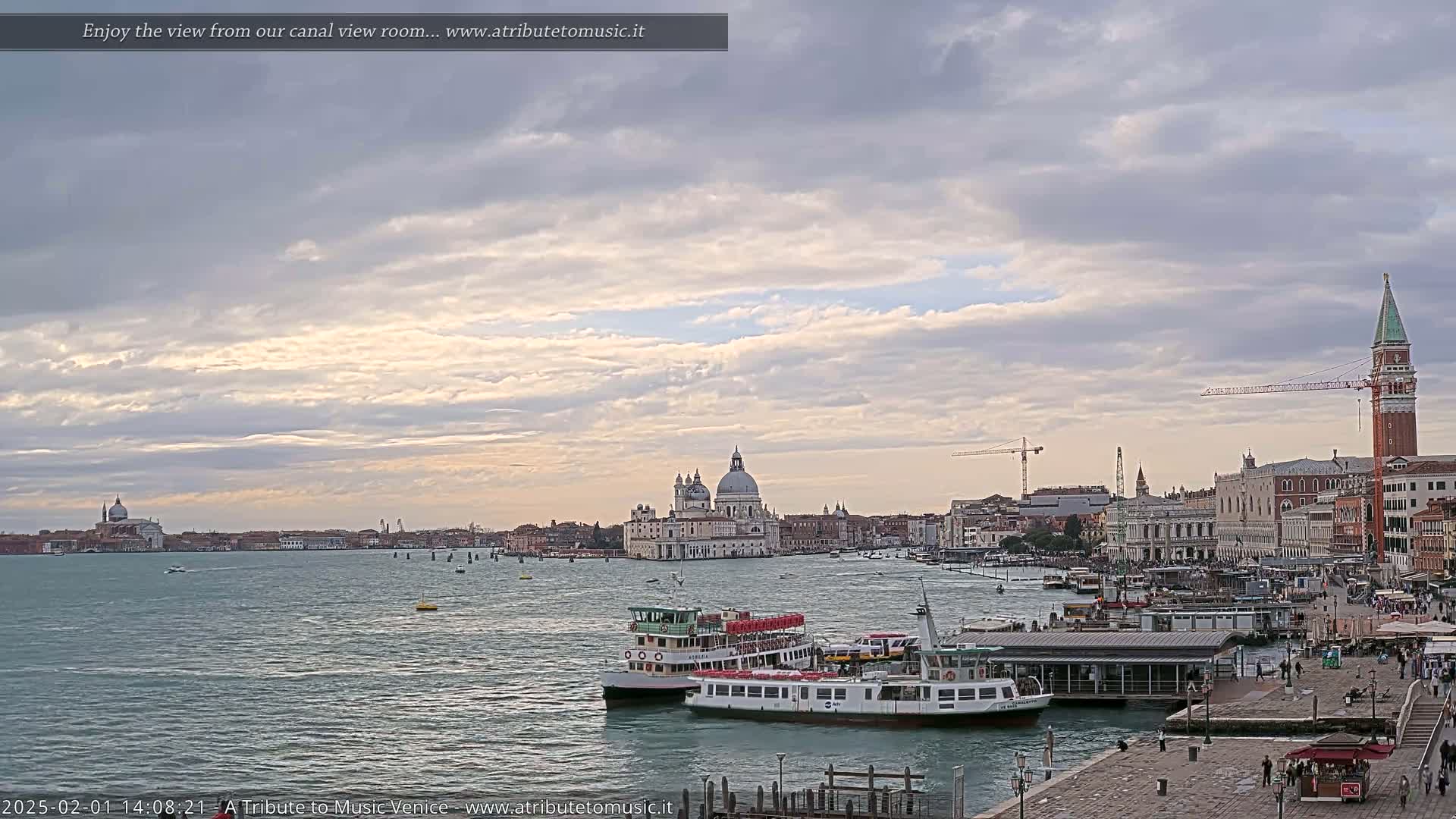 A wide shot of a Venetian canal shows several water buses near a dock, with the Basilica di Santa Maria della Salute and other buildings visible across the water under a cloudy sky.