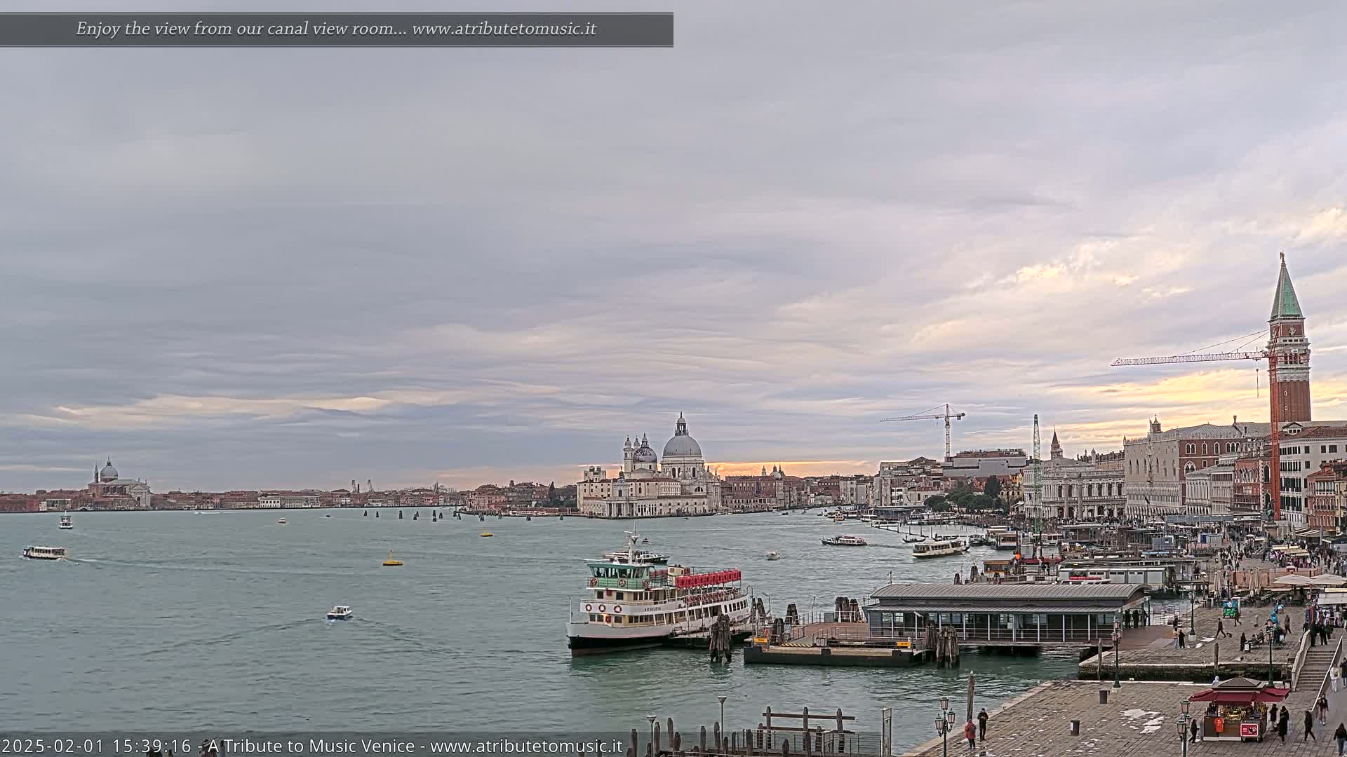 A wide shot of a Venetian canal shows a ferry docked near a building, with several boats on the water, and the city skyline featuring churches and a tall tower under a cloudy sky.