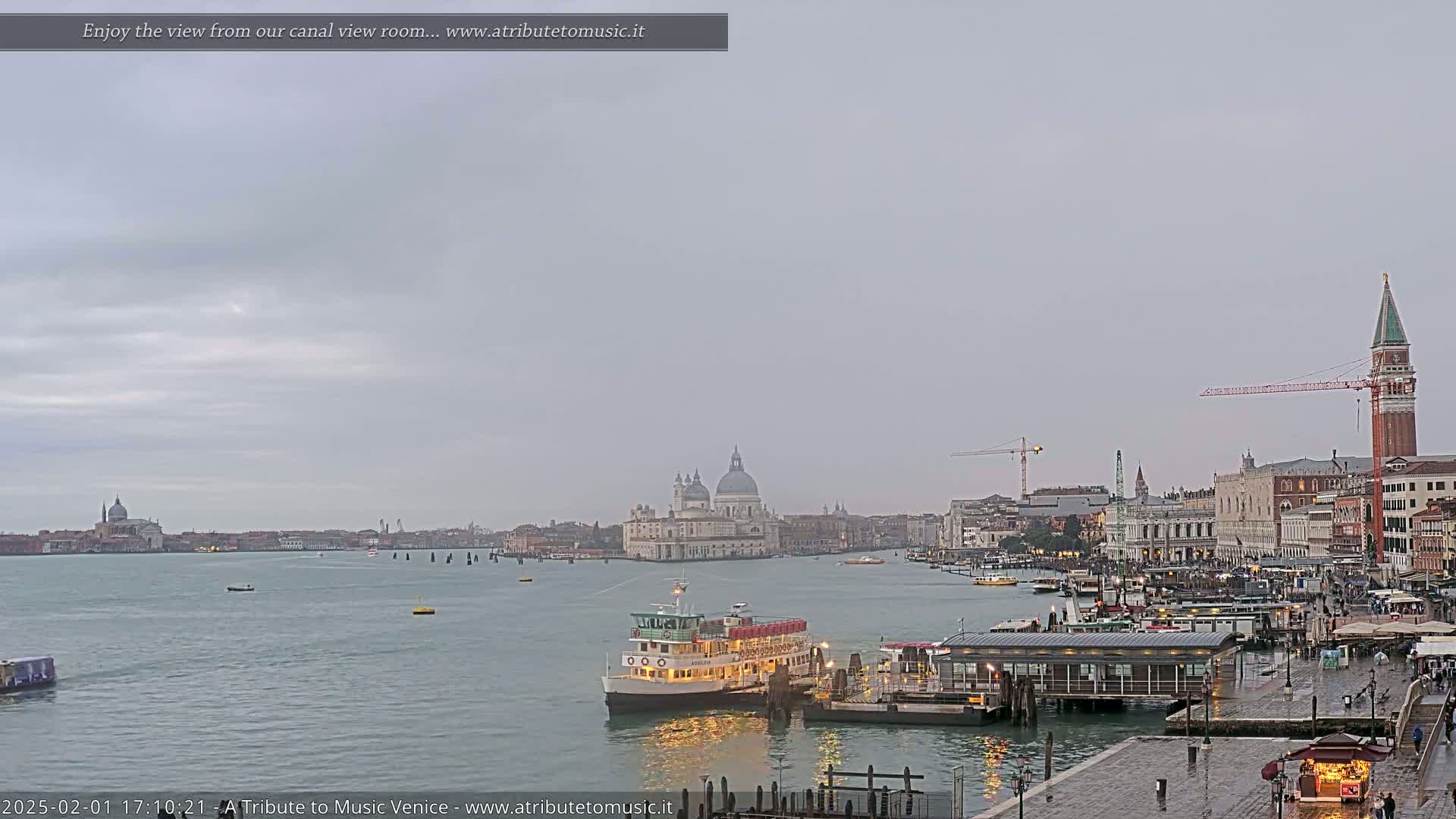 A ferry is docked at a waterfront station in Venice under an overcast sky, with the Basilica di Santa Maria della Salute visible in the distance.