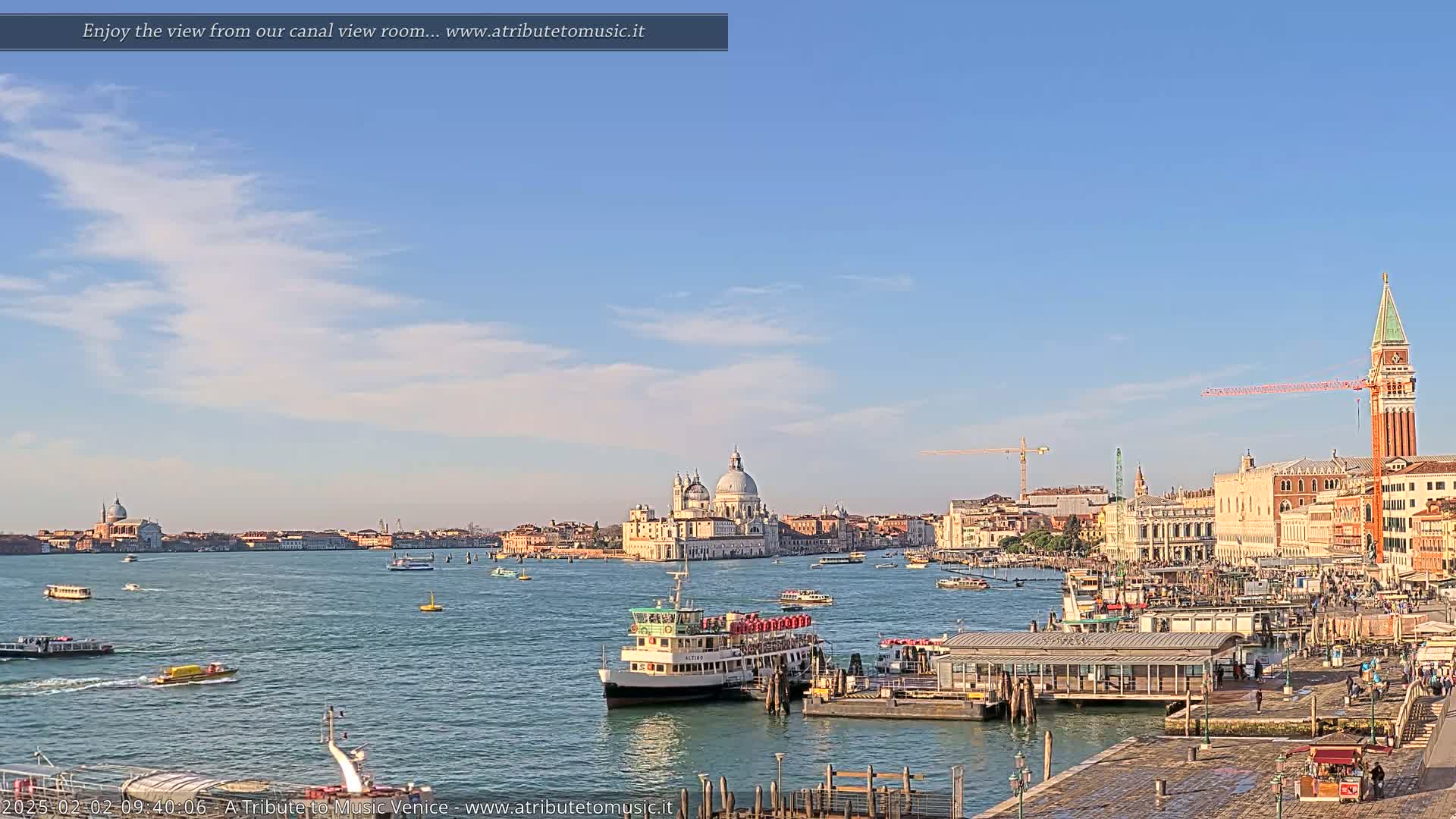 A sunny Venetian cityscape with a calm waterway filled with boats, showcasing a large church and the St. Mark's Campanile under a partly cloudy sky.