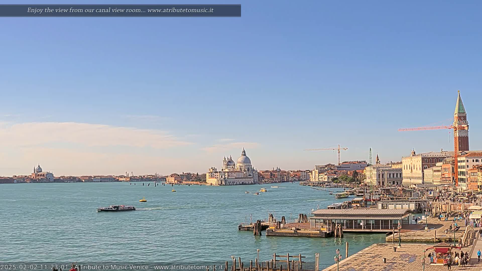 A clear, sunny day reveals a Venetian canal scene with boats, a waterfront building, and a distant view of the Basilica di Santa Maria della Salute and St. Mark’s Square.