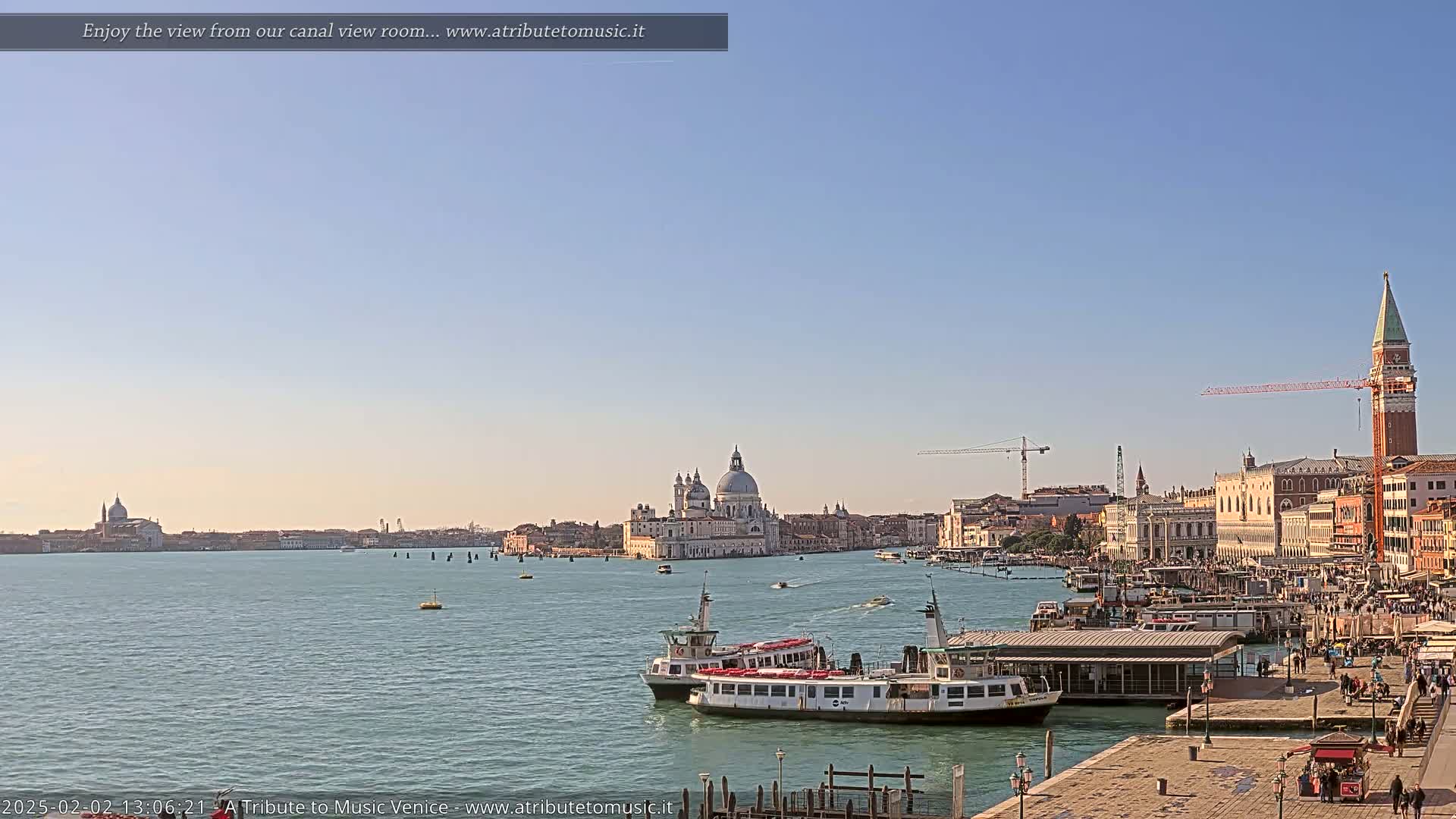 A sunny day in Venice shows a wide canal with several boats, a large church in the distance, and buildings along the waterfront.