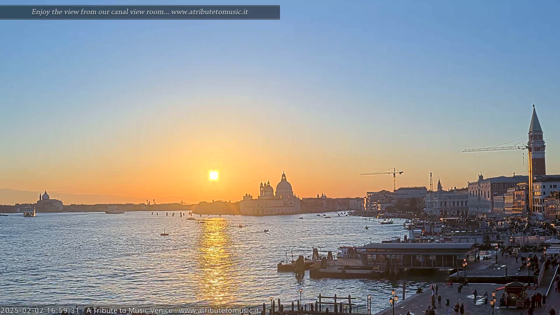 A calm Venetian canal at sunset, with the sun reflecting on the water, shows several boats and buildings along the waterfront, including a large domed church and a tall bell tower.