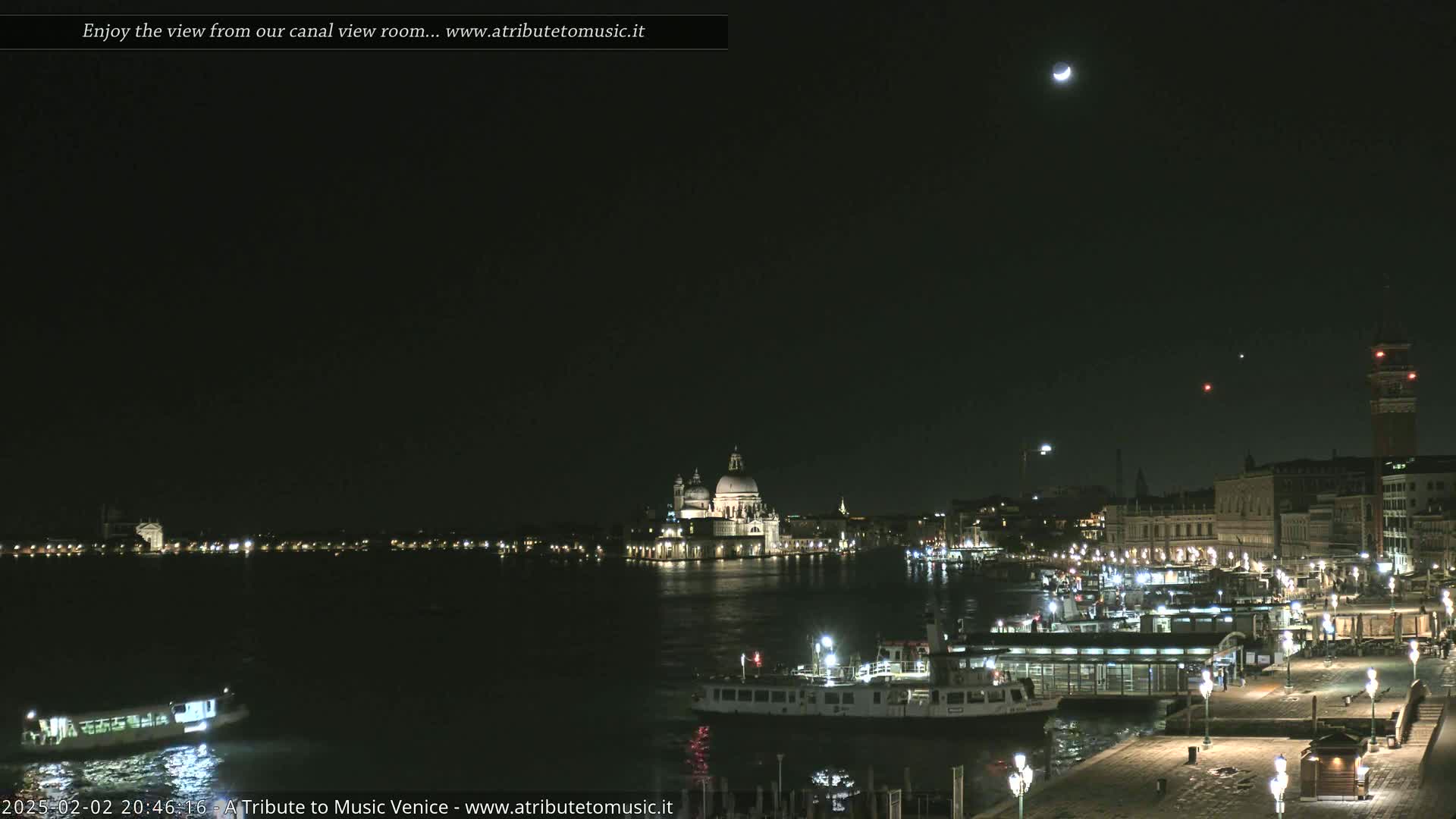 A nighttime view of a canal in Venice, showing boats, buildings, and a crescent moon in a clear sky.