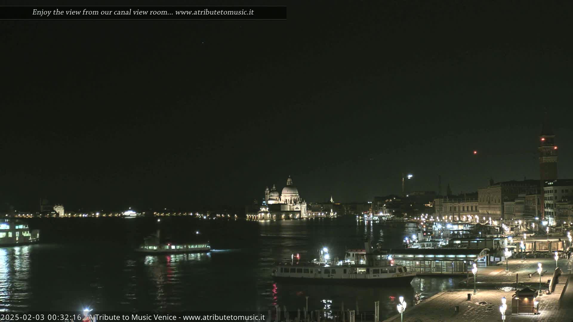 A nighttime view of a canal in Venice, Italy, shows several boats and illuminated buildings, including a large domed church, under a clear, dark sky.