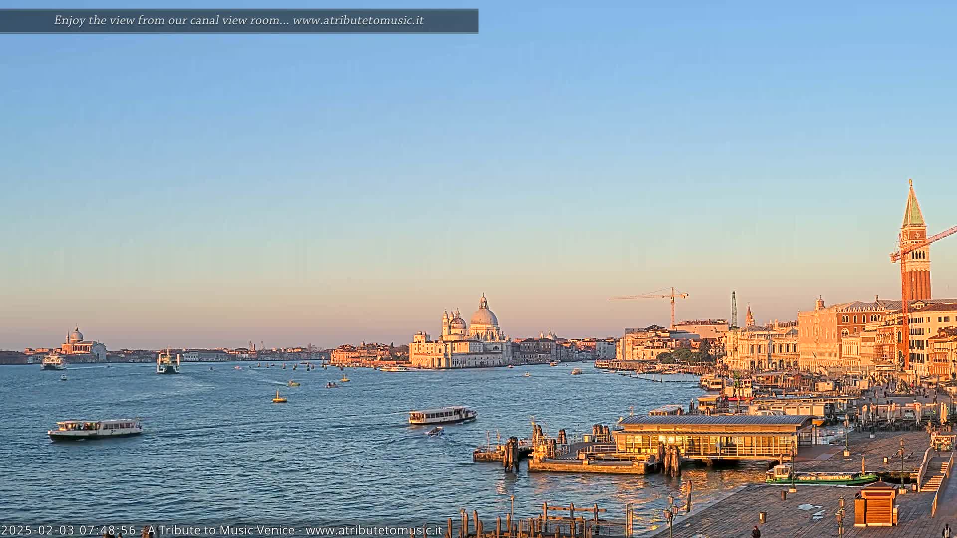 A tranquil Venetian canal scene at sunrise, showing a cityscape with boats, a church, and a bell tower under a clear, light-blue sky.