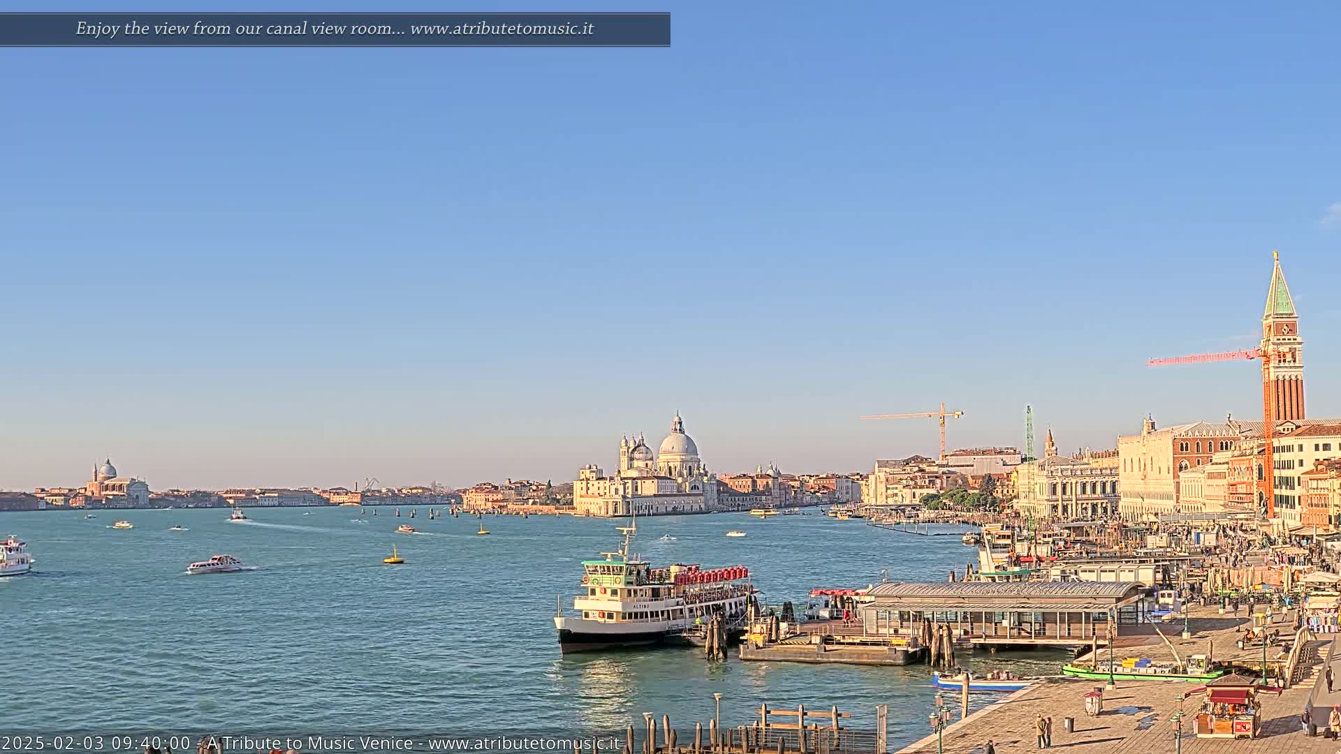 A sunny day in Venice shows a wide canal with boats, a ferry at a dock, and the city skyline including the Basilica di Santa Maria della Salute and St. Mark's Campanile in the distance.
