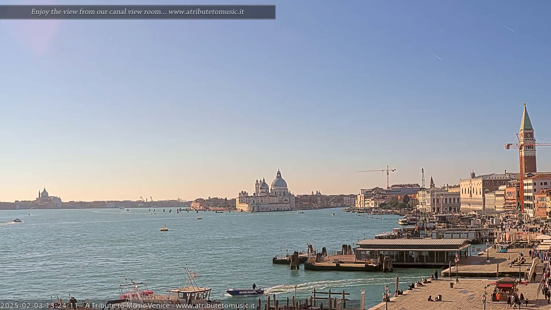 A wide shot of a Venetian canal on a clear, sunny day shows a cityscape with boats, buildings, and a large church in the distance.
