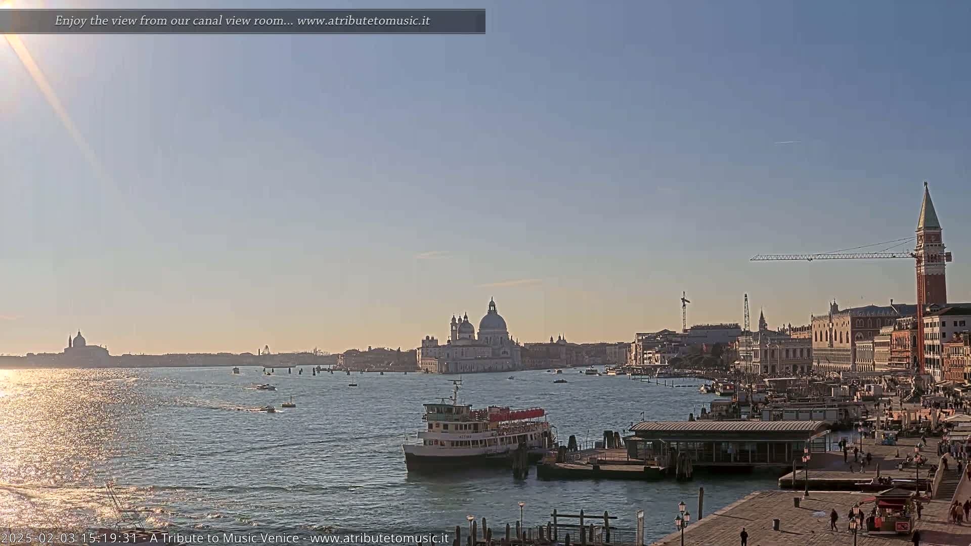 A sunny day in Venice shows a canal with boats and a view of the city skyline, including a large domed church and a tall bell tower.