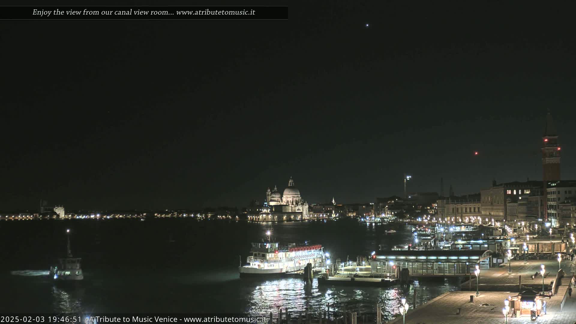 A nighttime view of a canal in Venice, showing boats docked near illuminated buildings and a large church in the distance under a clear, dark sky.