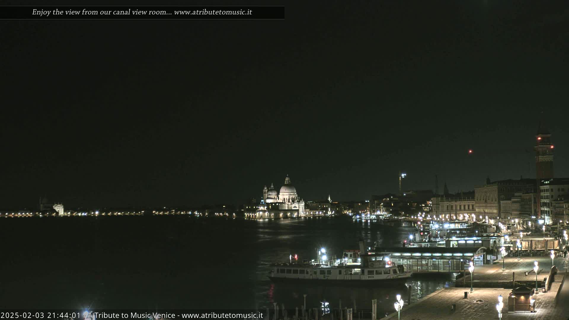 A nighttime view across a calm canal shows a city with illuminated buildings, boats, and a large domed structure.
