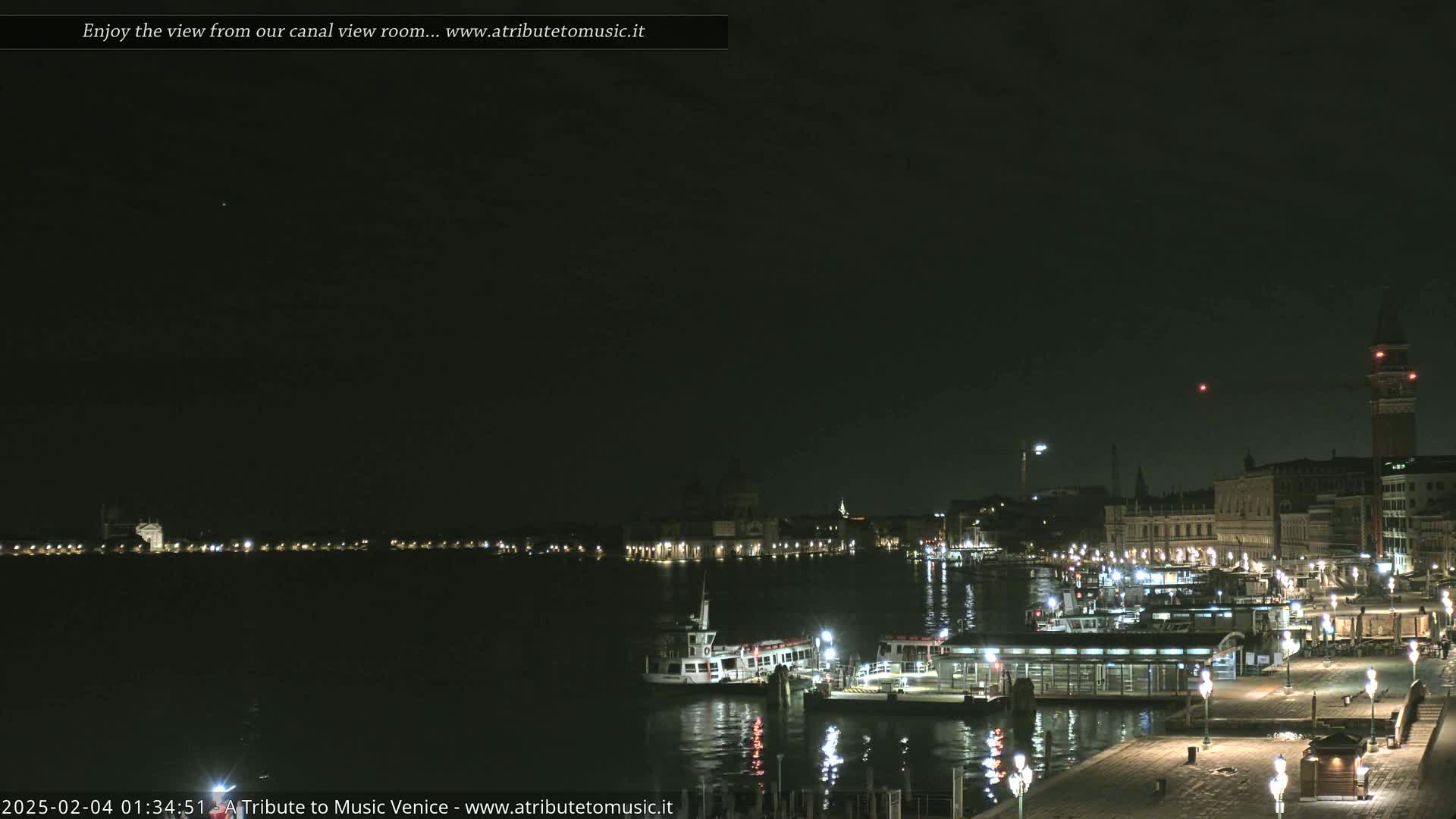 A nighttime view across a calm canal shows a city waterfront with buildings, boats, and docked ferries under a dark sky.
