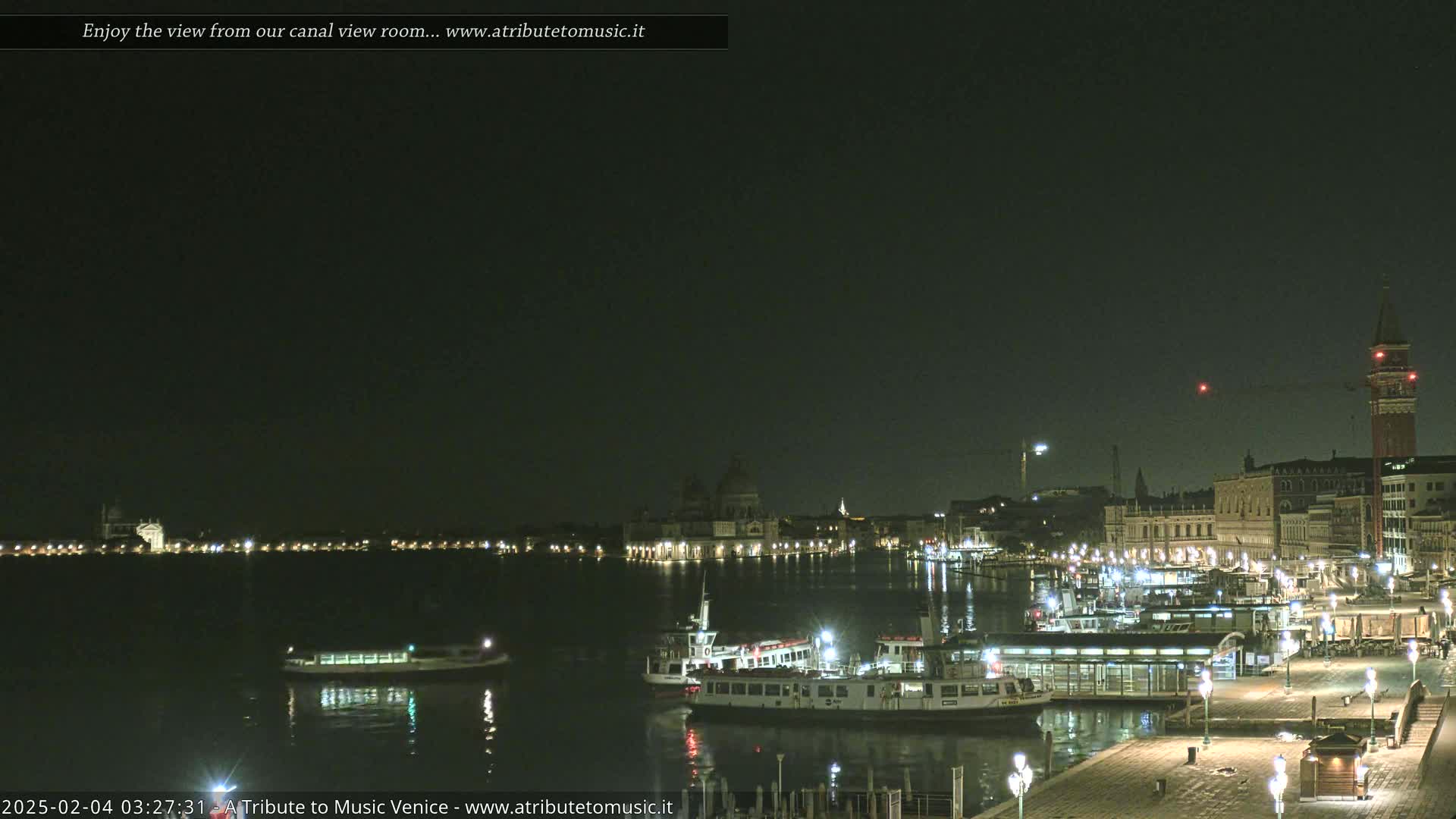 A nighttime view of a canal in Venice, Italy, shows illuminated buildings and several boats moored along a waterfront.