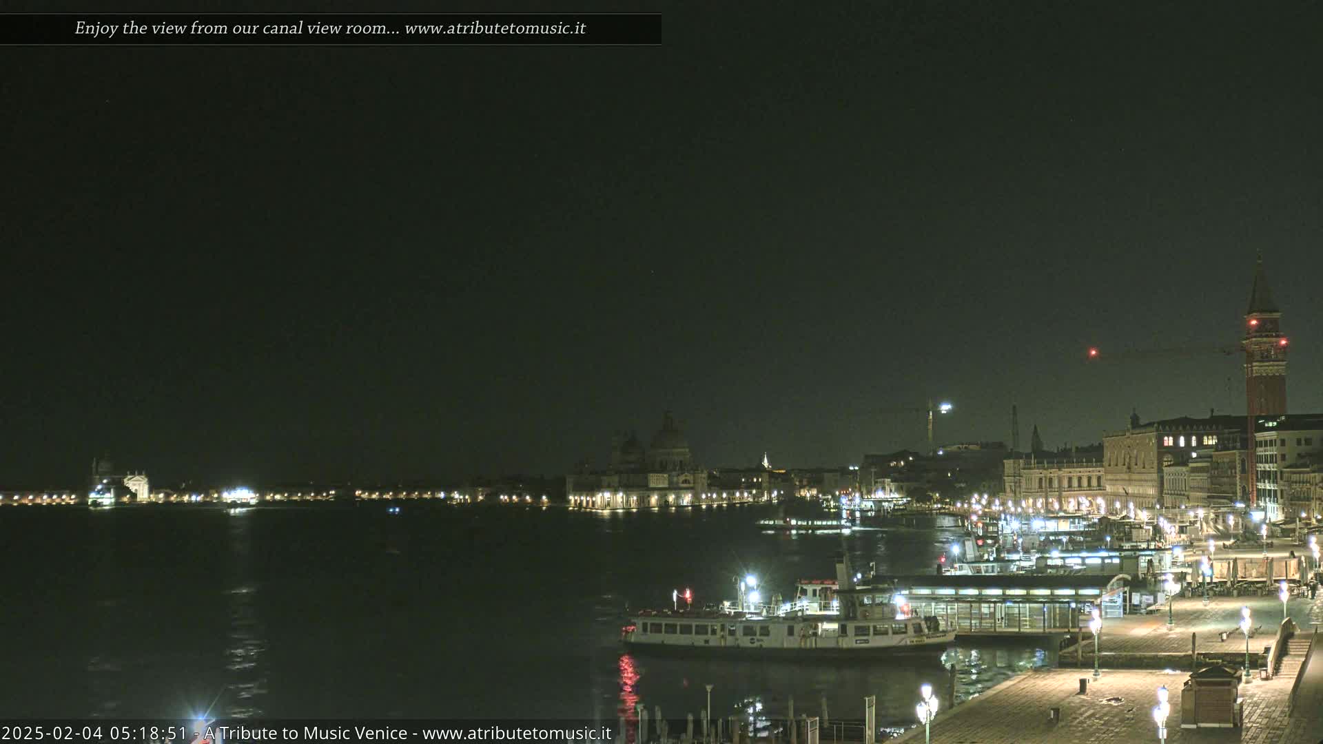 A nighttime view across a calm body of water shows illuminated buildings and boats along a waterfront.