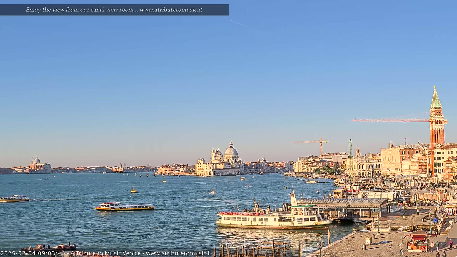 A sunny Venetian cityscape is viewed across a calm canal, featuring a large church, several boats, and the San Giorgio Maggiore bell tower.