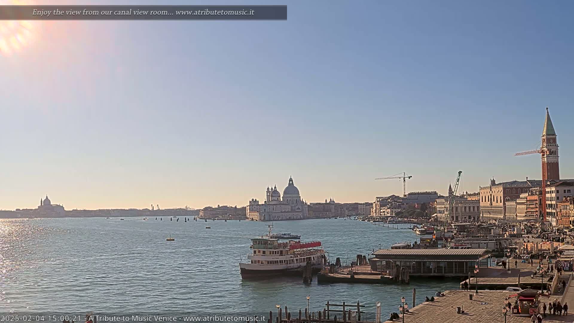 A Venetian waterway is shown, with a ferry docked near a city waterfront, under a clear, sunny sky;  in the distance, the domes of a church and a tall bell tower are visible.