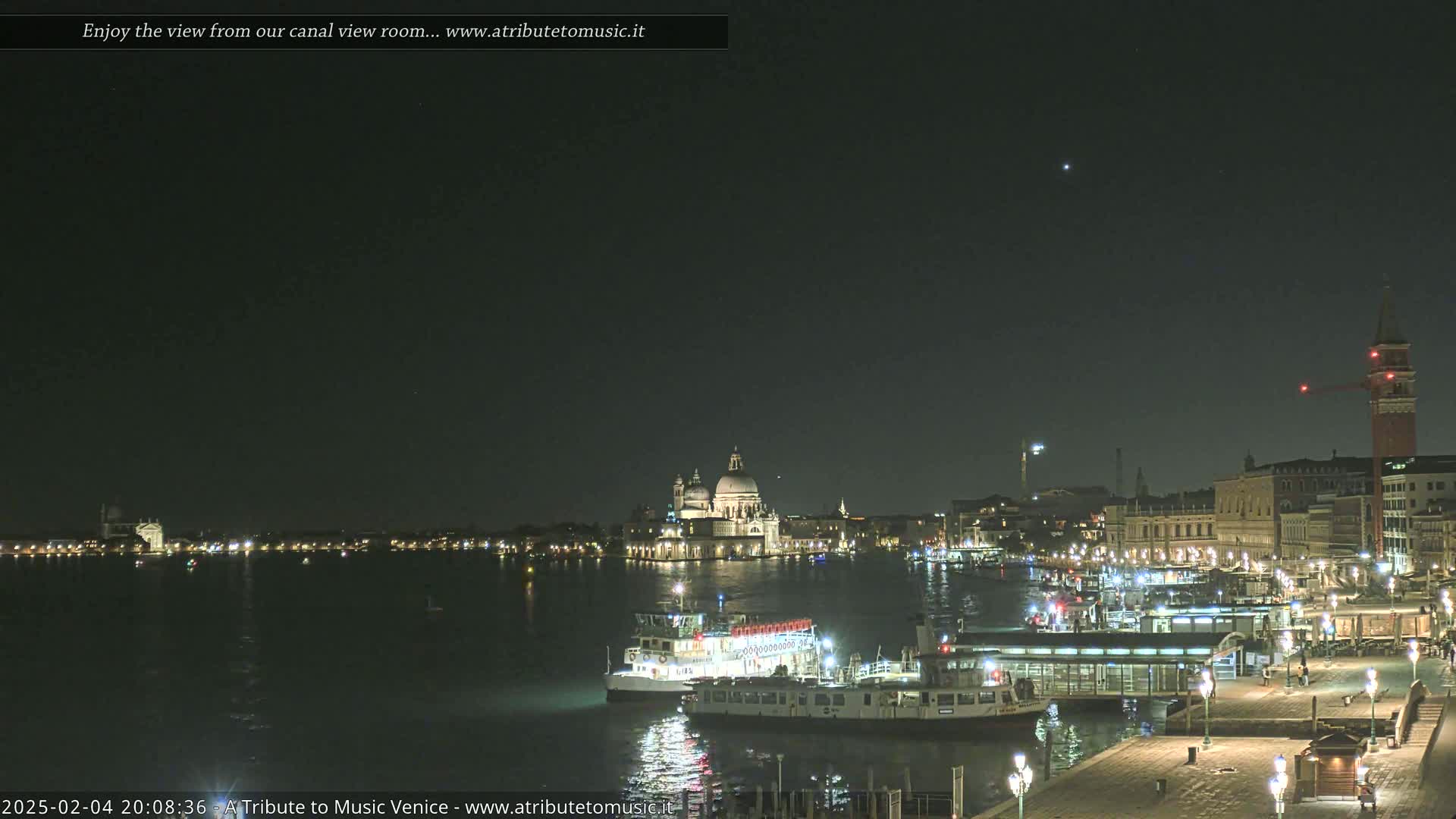 A nighttime view of a canal in Venice, Italy, shows several boats docked near buildings along the waterfront, with the Basilica di Santa Maria della Salute illuminated in the distance under a clear, dark sky.