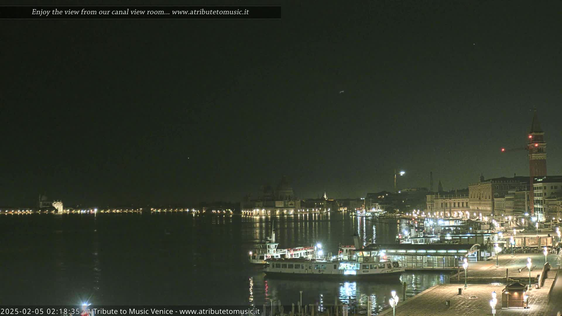 A nighttime view across a calm canal shows illuminated buildings and several boats docked along a waterfront, under a dark, clear sky.