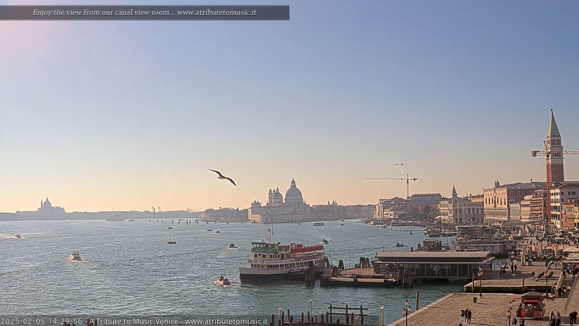 A clear, sunny day in Venice shows a wide canal with boats and a city skyline including a large church and a tall bell tower.