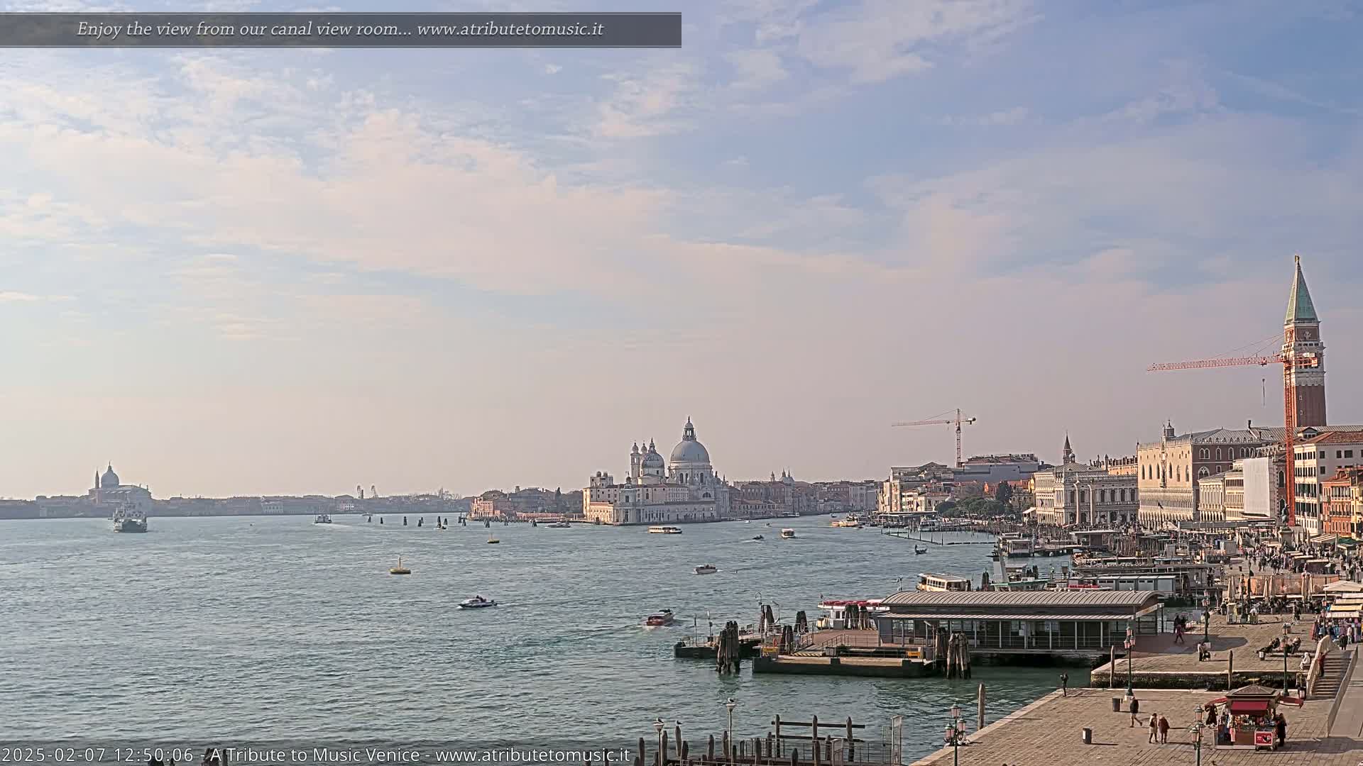 A wide shot of a Venetian canal on a partly cloudy day shows a large body of water with boats, a city skyline including a large church, and a waterfront area with people.