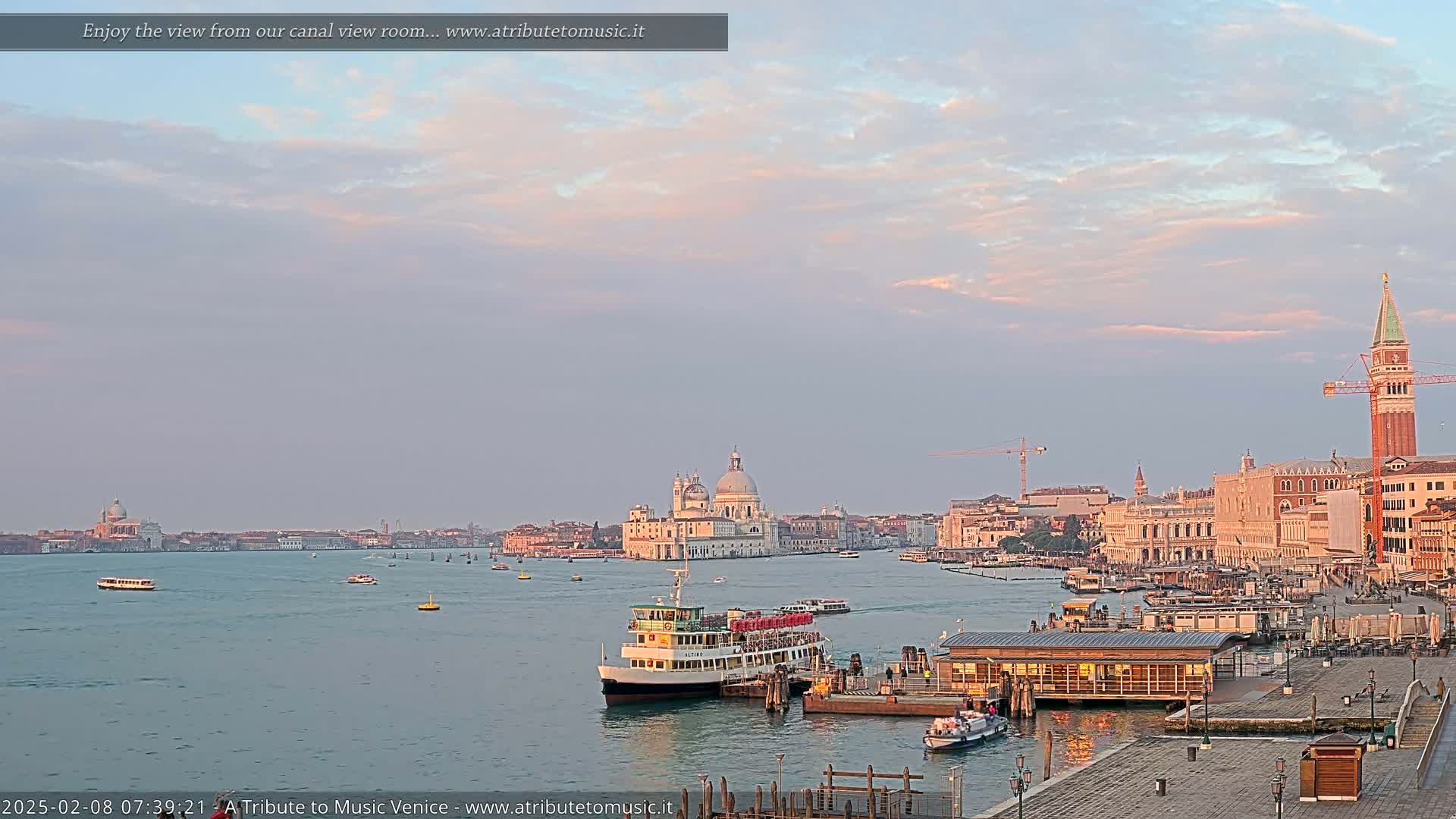 A calm Venetian canal at sunrise, showing a ferry docked near buildings and a church in the distance under a pale pink and cloudy sky.