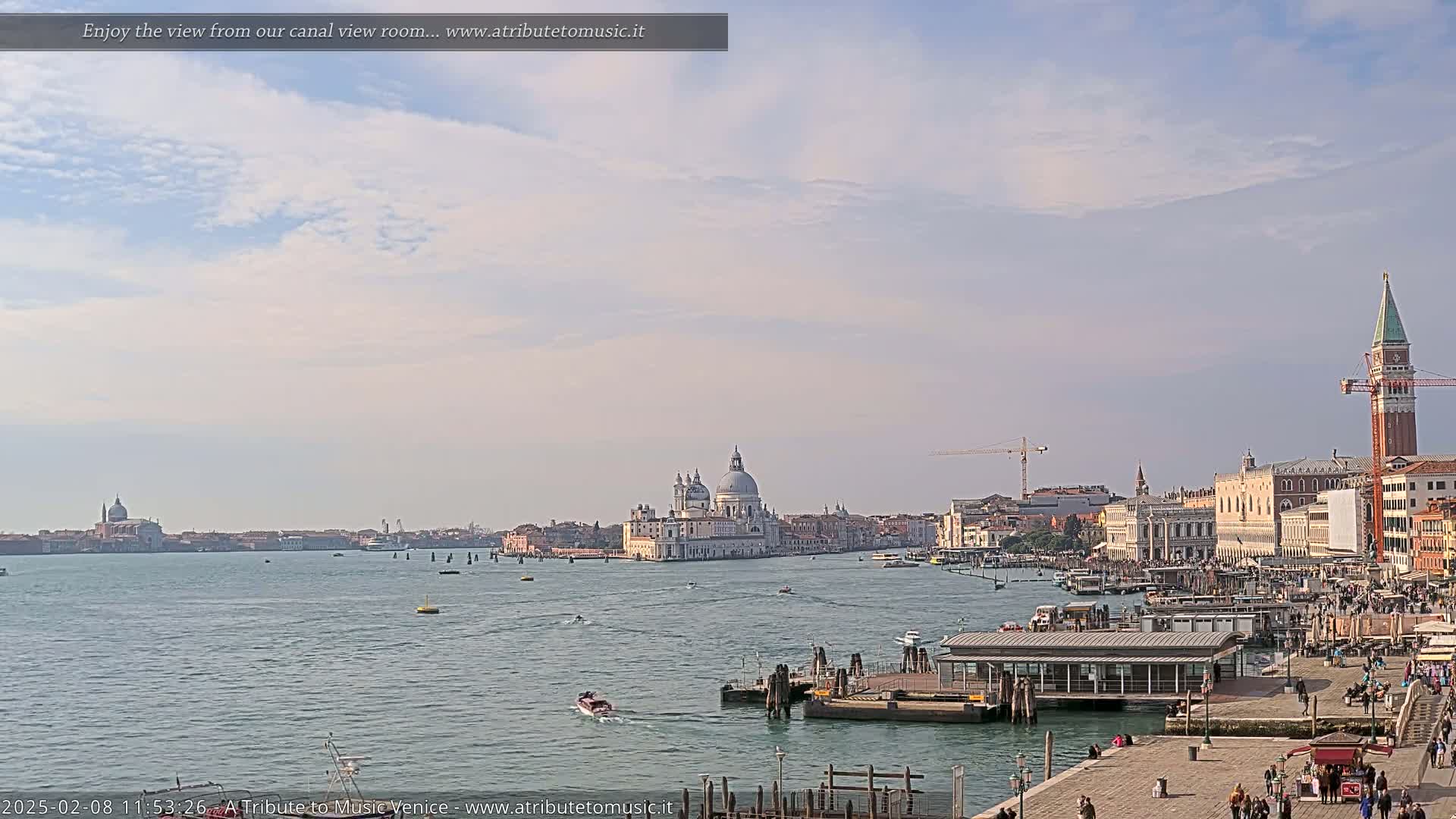 A wide, long shot of a waterway shows a city with buildings, a church dome, and a tall tower under a mostly clear, lightly cloudy sky.
