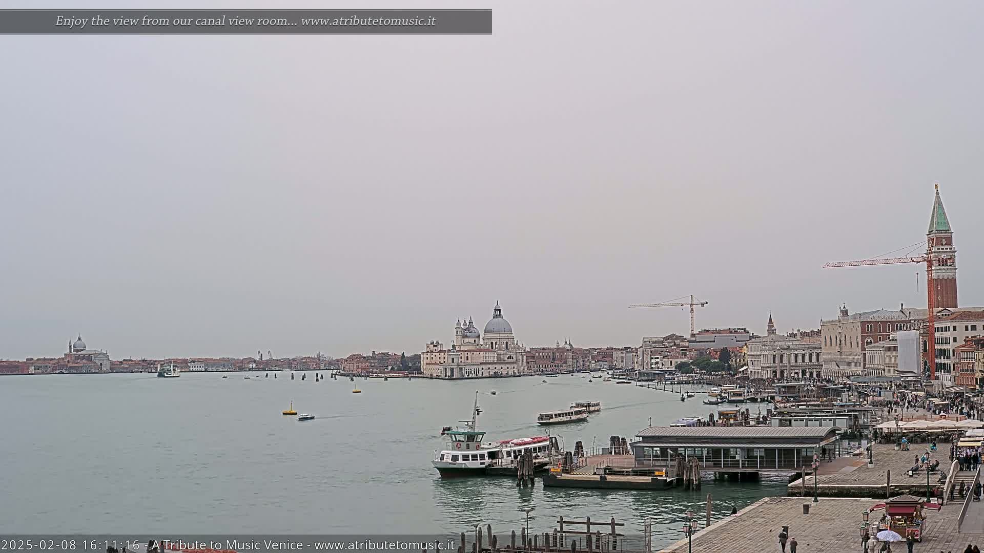 A wide shot of a Venetian canal on an overcast day shows a waterway with several boats, including a ferry, and buildings lining the waterfront, featuring a prominent domed church.