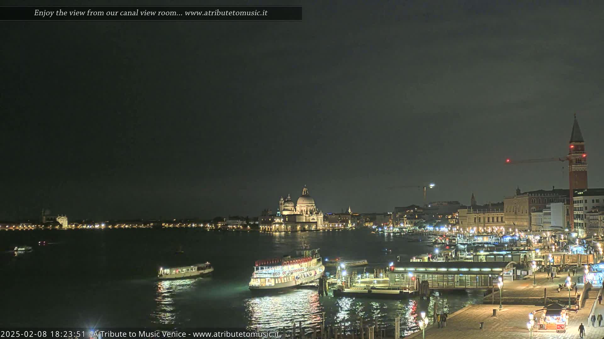 A nighttime view of a Venetian canal with boats, illuminated buildings, and a large church in the distance under a dark sky.