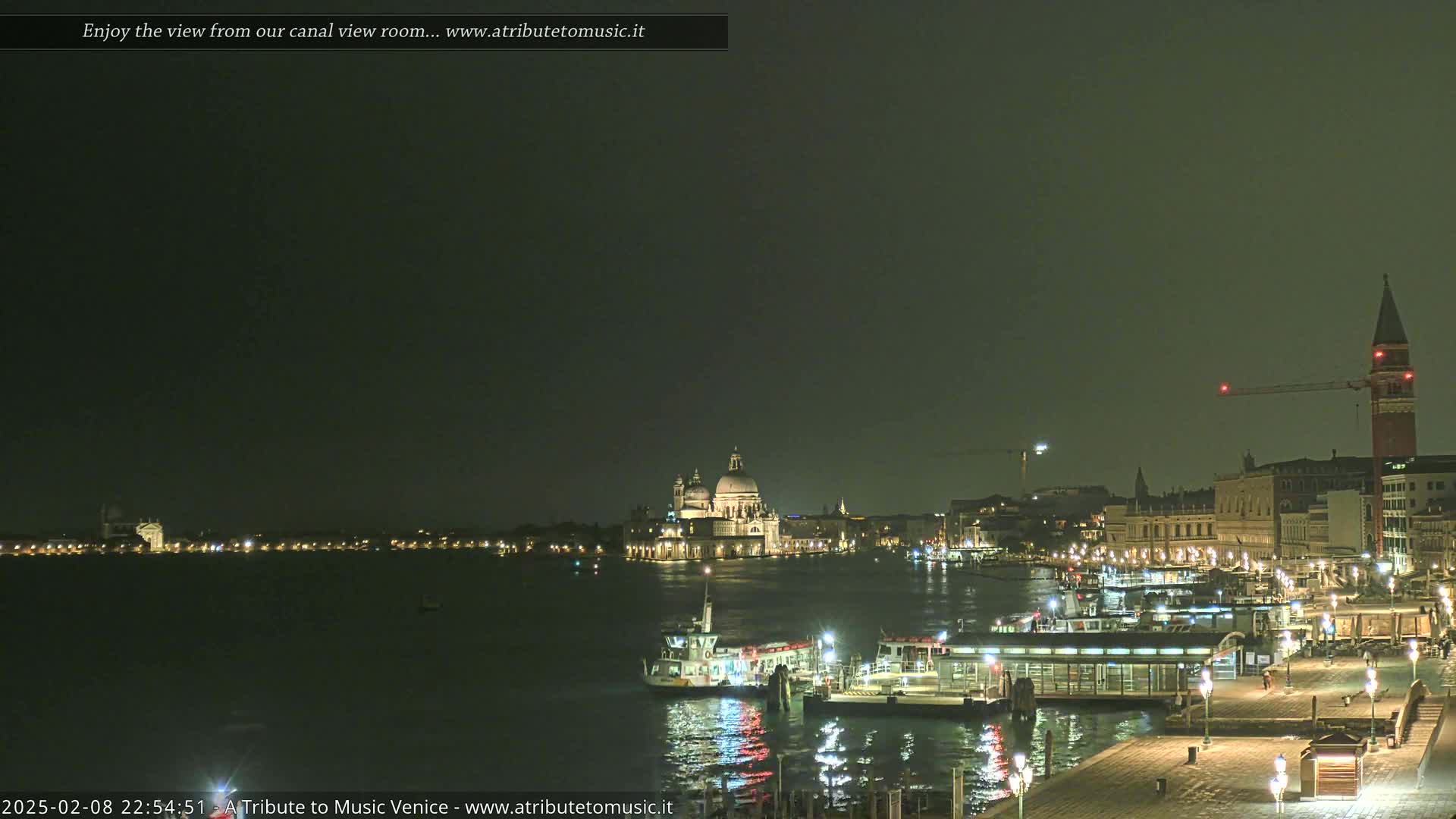 A nighttime view of a canal in Venice, Italy, shows illuminated buildings, boats, and a pier under a dark, overcast sky.