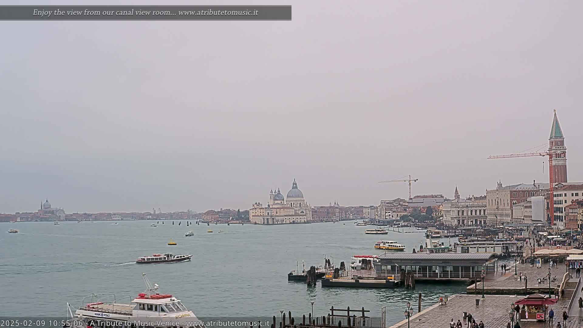 A wide shot of a Venetian canal on an overcast day shows several boats on the water with the Basilica di Santa Maria della Salute and other buildings visible in the distance.