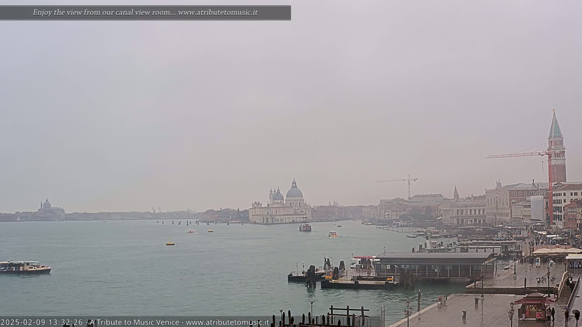 A hazy, rainy view of Venice, Italy shows a wide canal with boats, buildings along the waterfront, and the Basilica di Santa Maria della Salute in the distance.