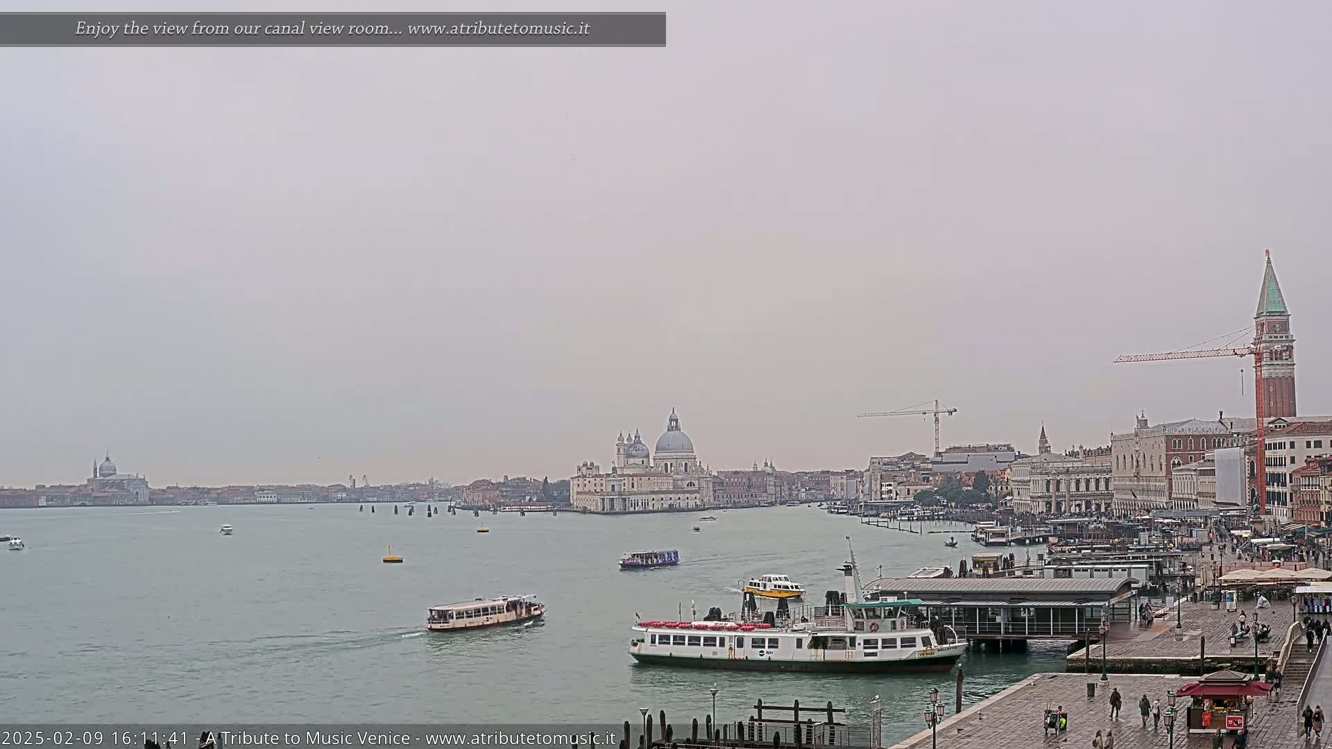 A wide shot of a Venetian canal on an overcast day shows several water taxis and a larger ferry, with the Basilica di Santa Maria della Salute and St. Mark's Campanile visible in the distance.