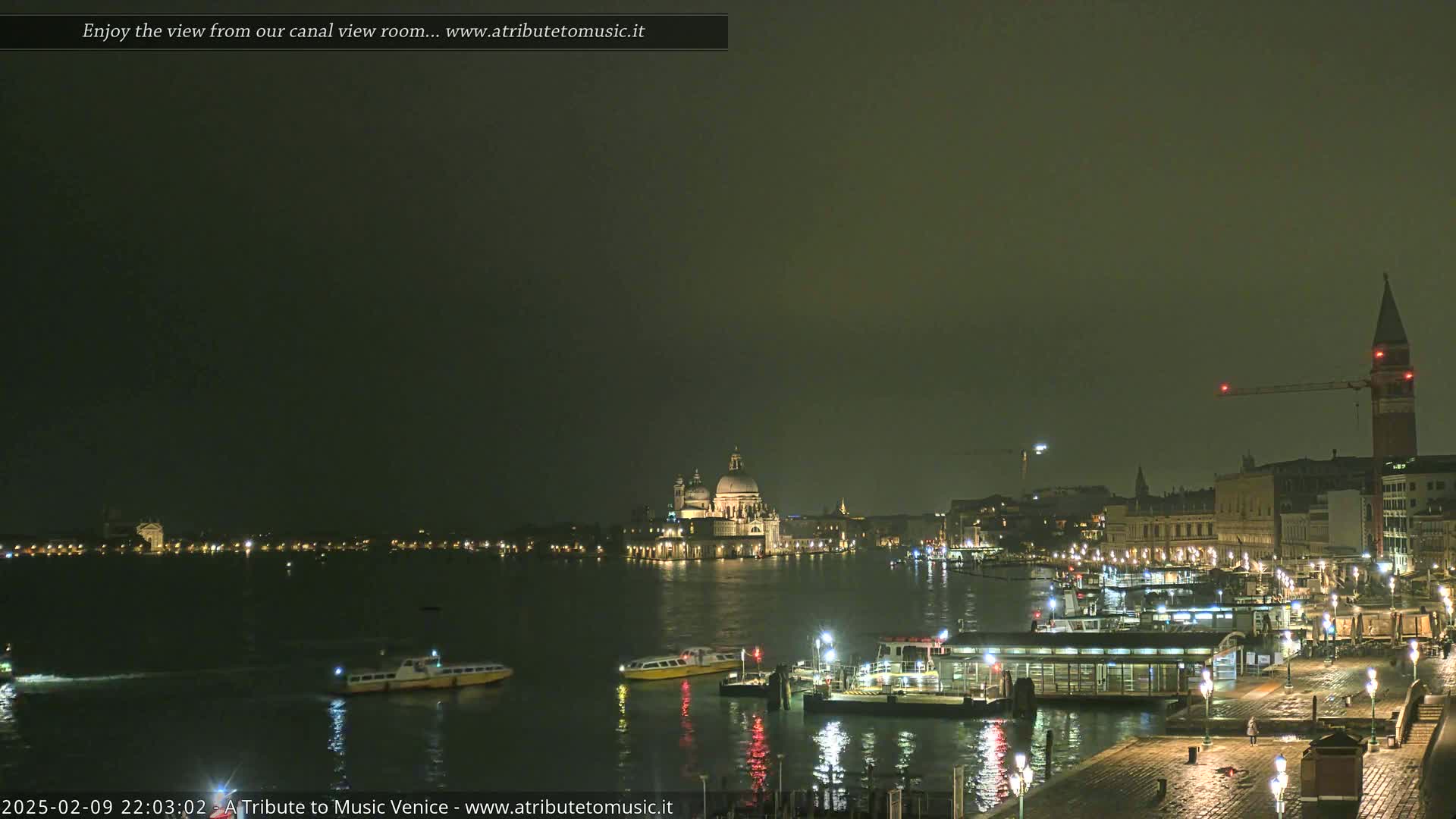 A nighttime view of a canal in Venice, Italy, shows several boats and buildings illuminated under a dark, overcast sky.