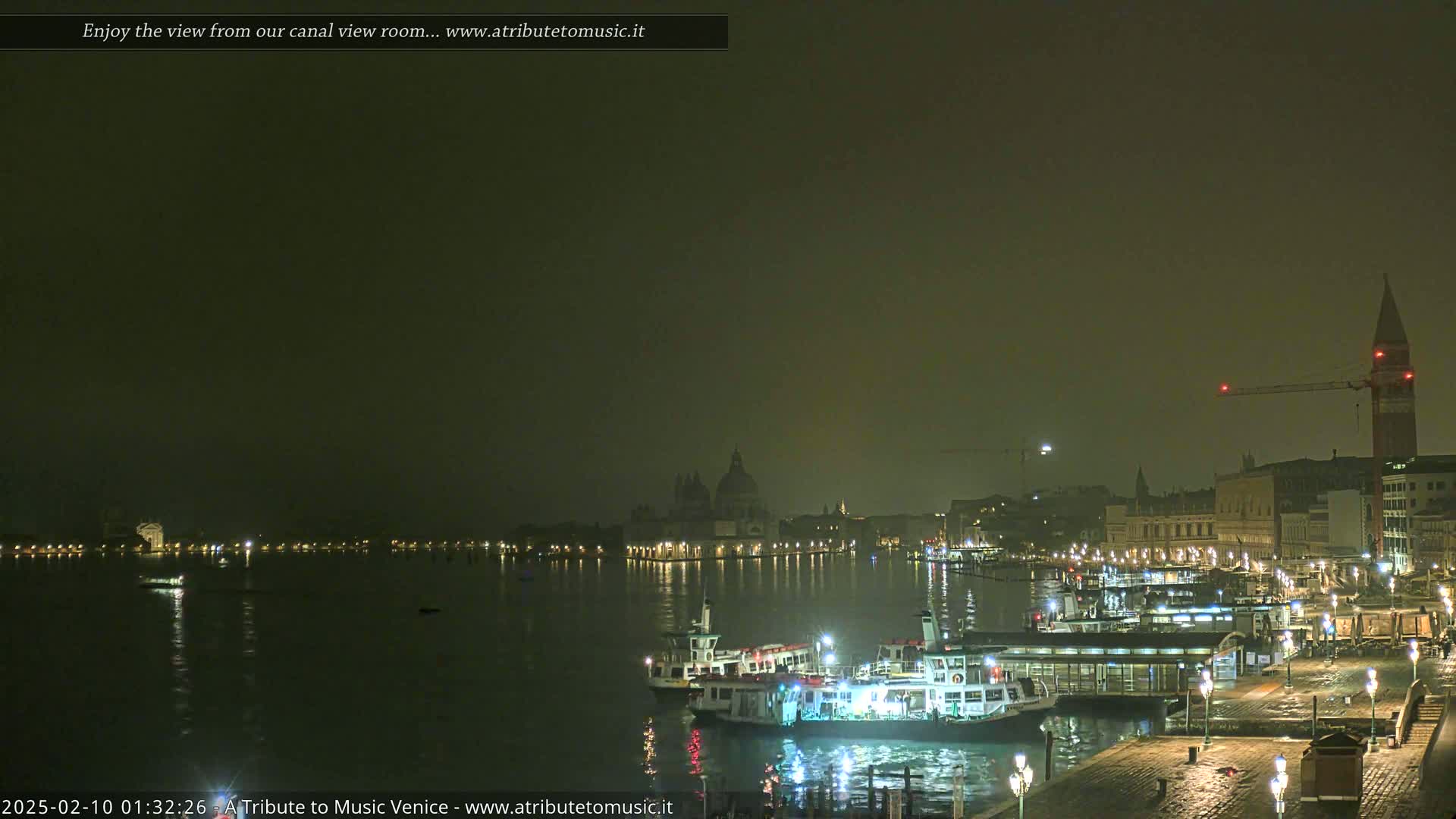 A nighttime view of a calm Venetian canal, showing several boats docked near illuminated buildings, with a hazy atmosphere.